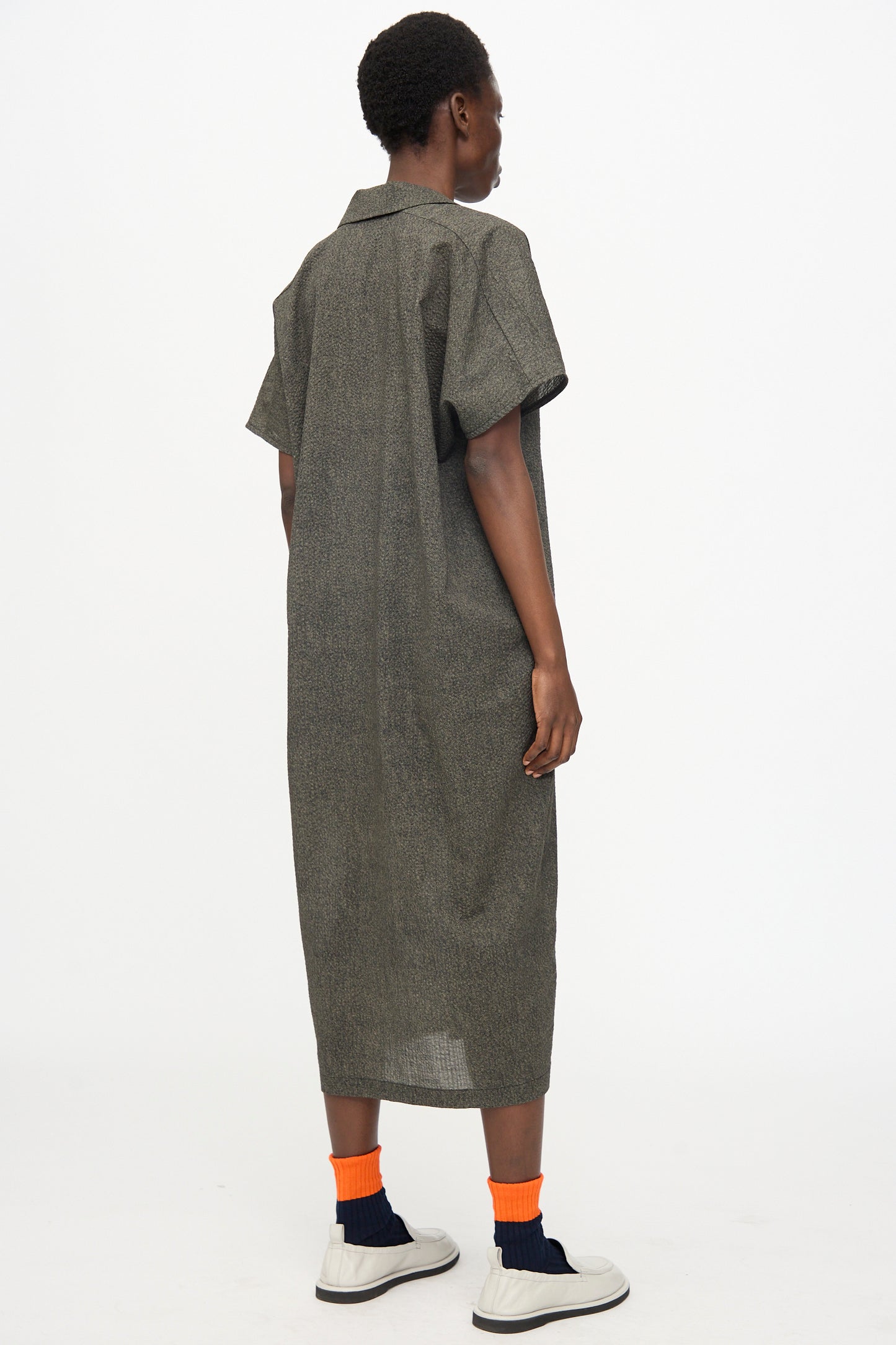 A person faces away from the camera, wearing the Black Crane Kite Dress with Micro Wave Texture in Steel, a relaxed fit, short sleeve dress in textured gray Japanese cotton, paired with white sneakers and orange socks.