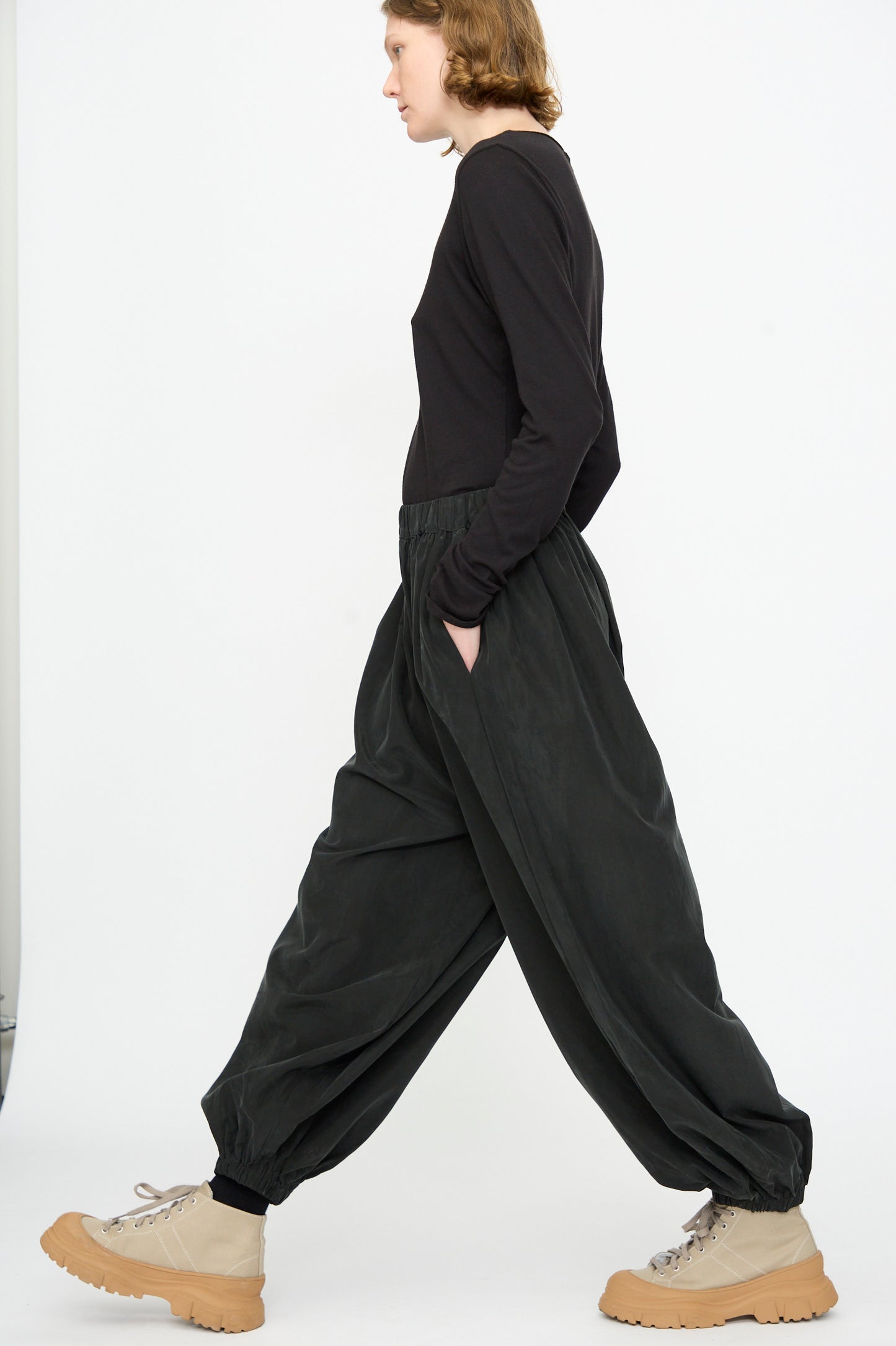 A person in profile view wears a black long-sleeve shirt, Black Crane Lantern Pants in Black—crafted from a comfortable cotton cupro blend with a loose, curved leg and elasticated waistband—and beige chunky boots, walking against a plain white background for an elevated everyday look.