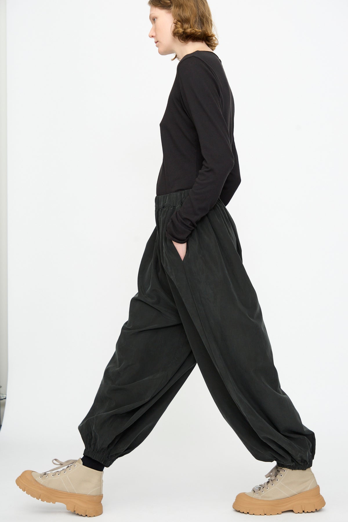 A person in profile view wears a black long-sleeve shirt, Black Crane Lantern Pants in Black—crafted from a comfortable cotton cupro blend with a loose, curved leg and elasticated waistband—and beige chunky boots, walking against a plain white background for an elevated everyday look.