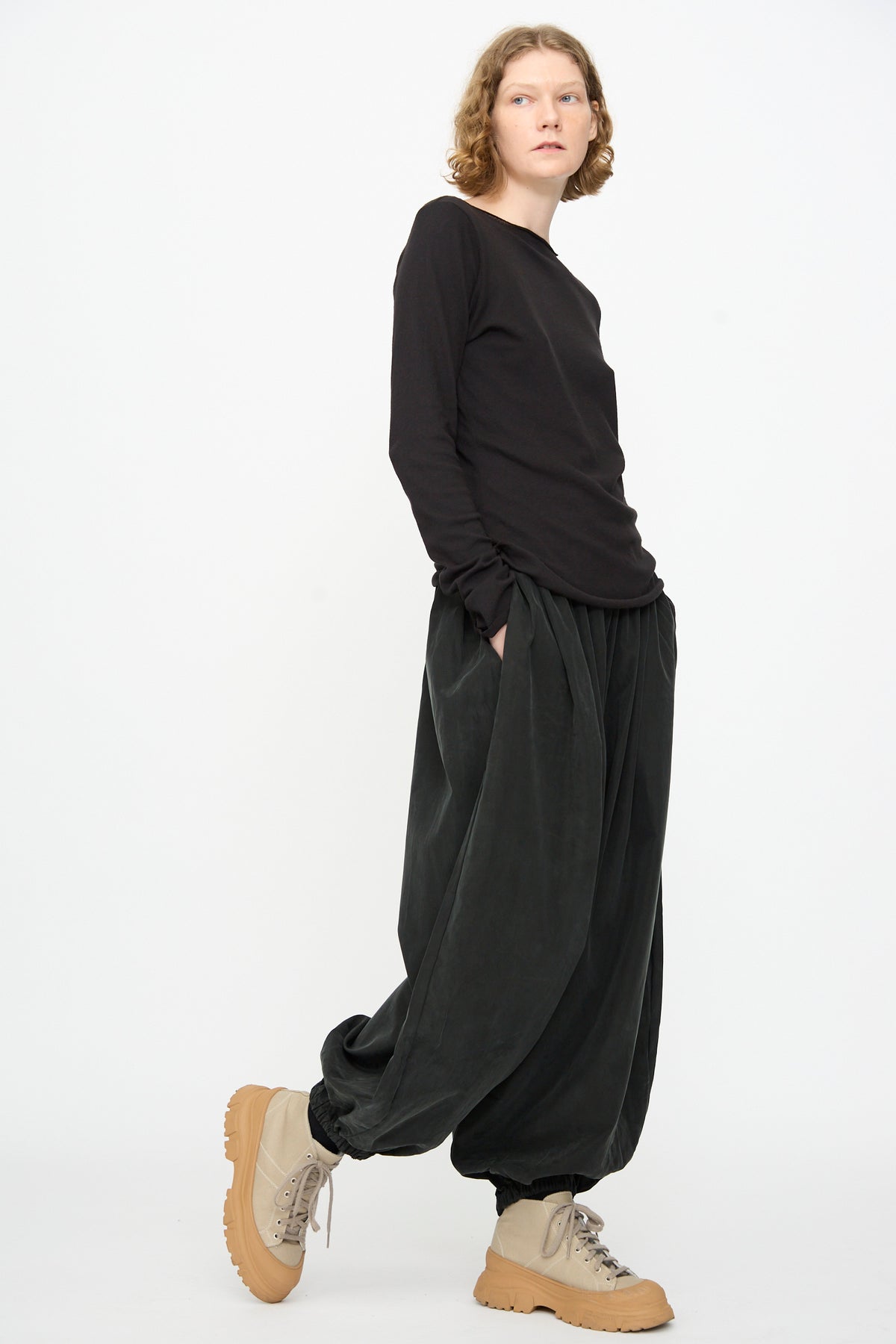A person wearing a loose black long-sleeve shirt and Black Crane’s Lantern Pants in Black—featuring a voluminous silhouette, cotton-cupro blend, elasticated waistband, and curved leg—paired with beige chunky shoes, stands against a plain white background.