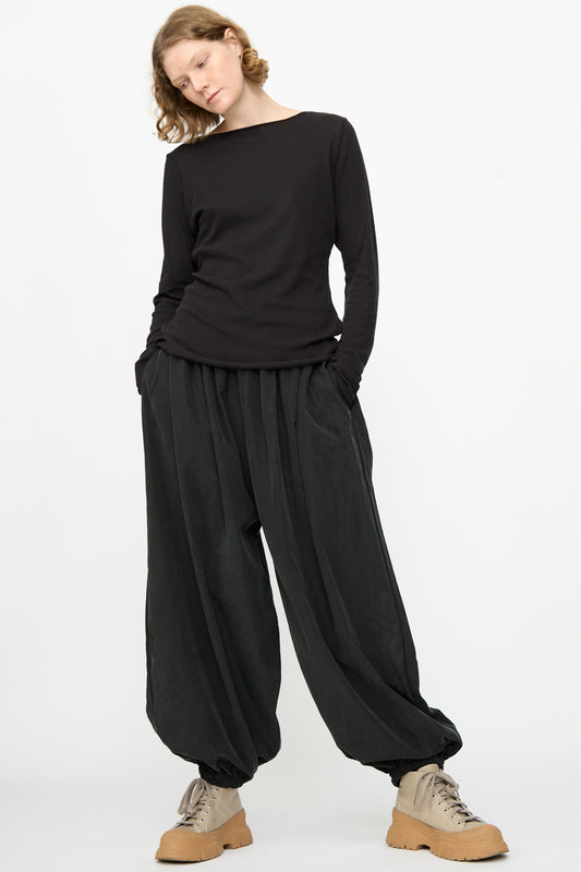 A person is wearing a loose black long-sleeve top, Black Crane Lantern Pants in Black featuring a wide curved leg and elasticated waistband in a soft cotton cupro blend, and beige lace-up boots, standing against a plain white background.