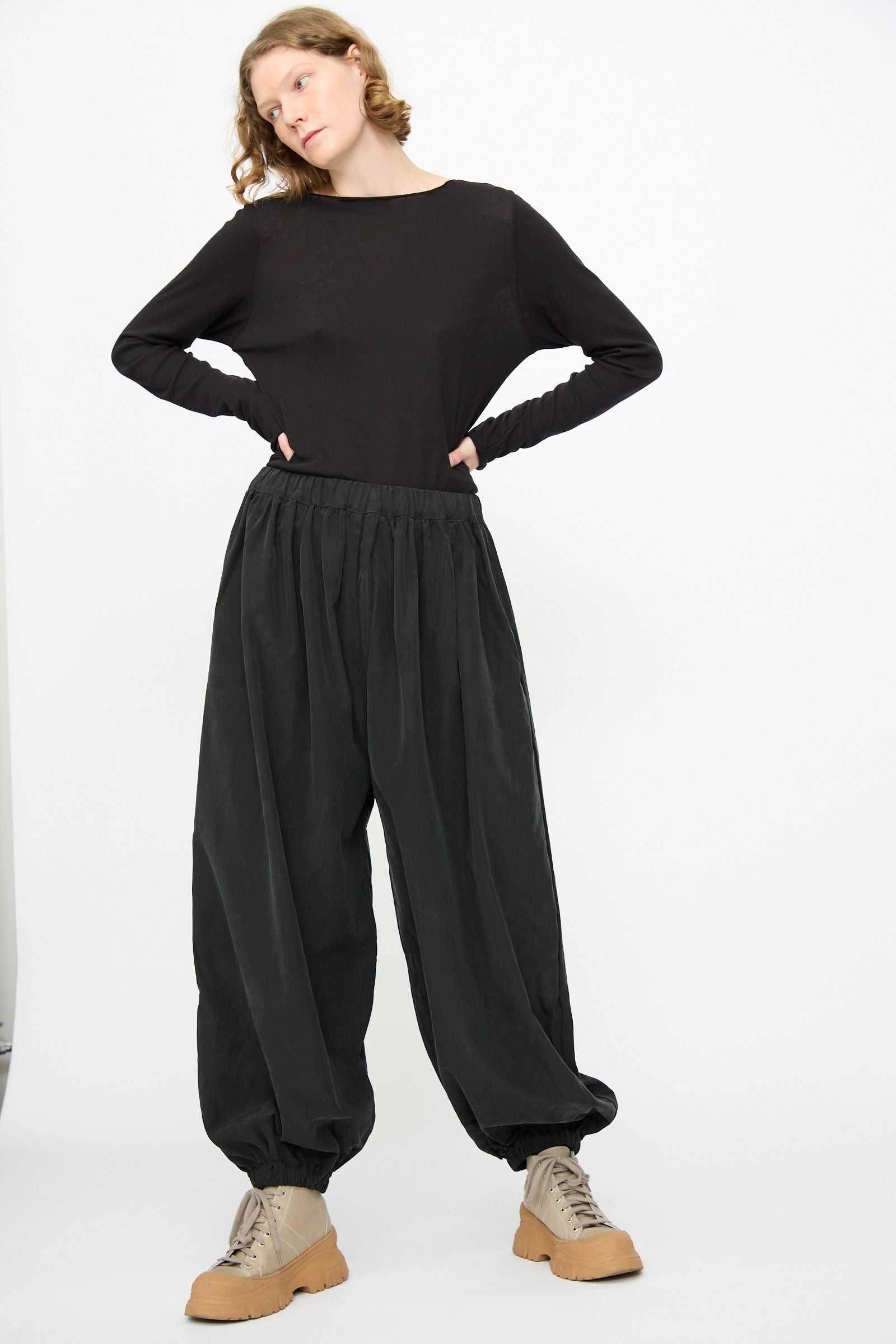 A person stands against a white background, wearing a black long-sleeve shirt, Black Crane Lantern Pants in Black featuring a loose curved leg and elasticated waistband crafted from a soft cotton cupro blend, paired with tan lace-up shoes.