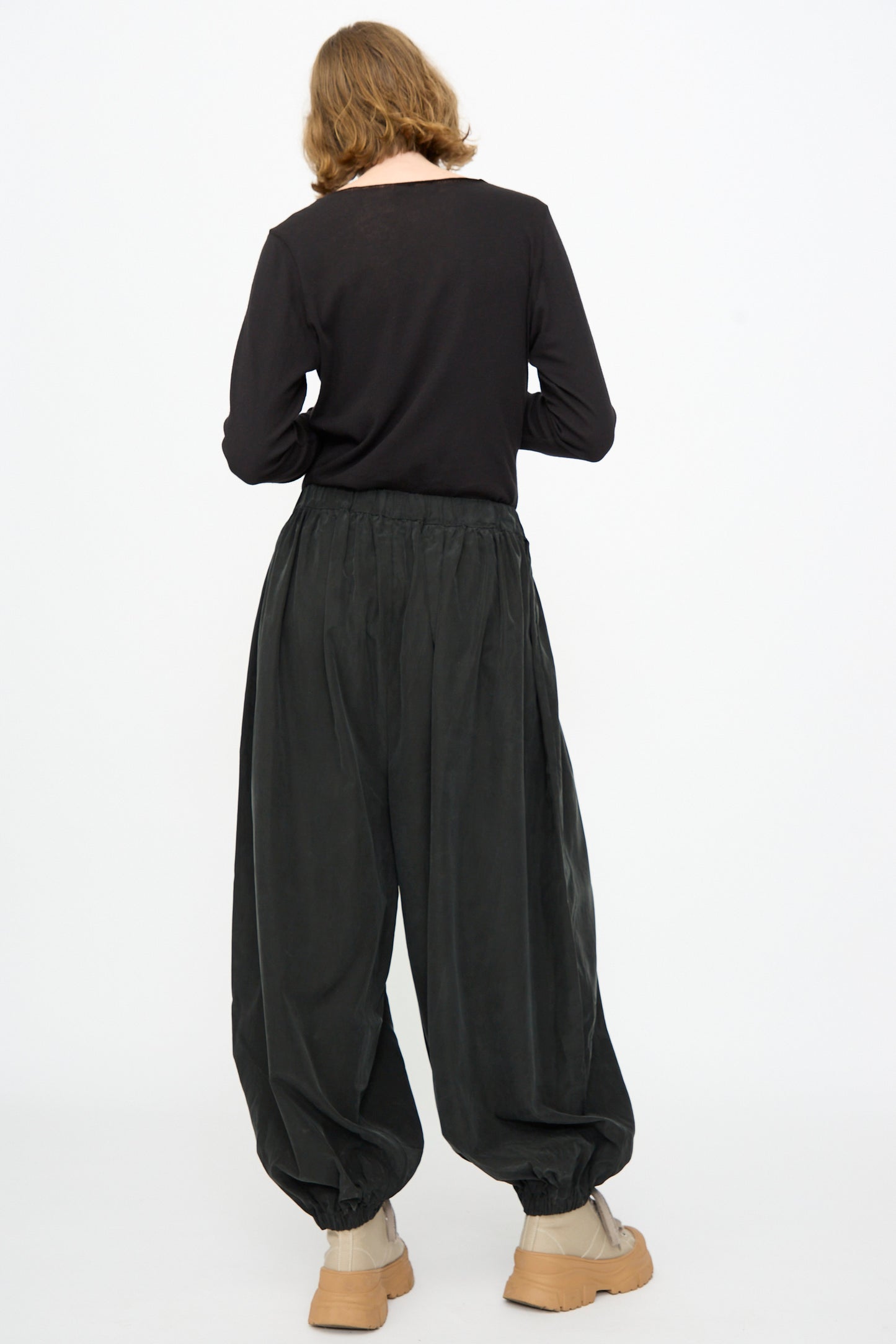 A person stands facing away against a plain white background, wearing Black Crane’s Lantern Pants in Black—a loose silhouette with an elasticated waistband and gathered ankles, crafted from a cotton cupro blend—paired with a black long-sleeve shirt and beige ankle boots.