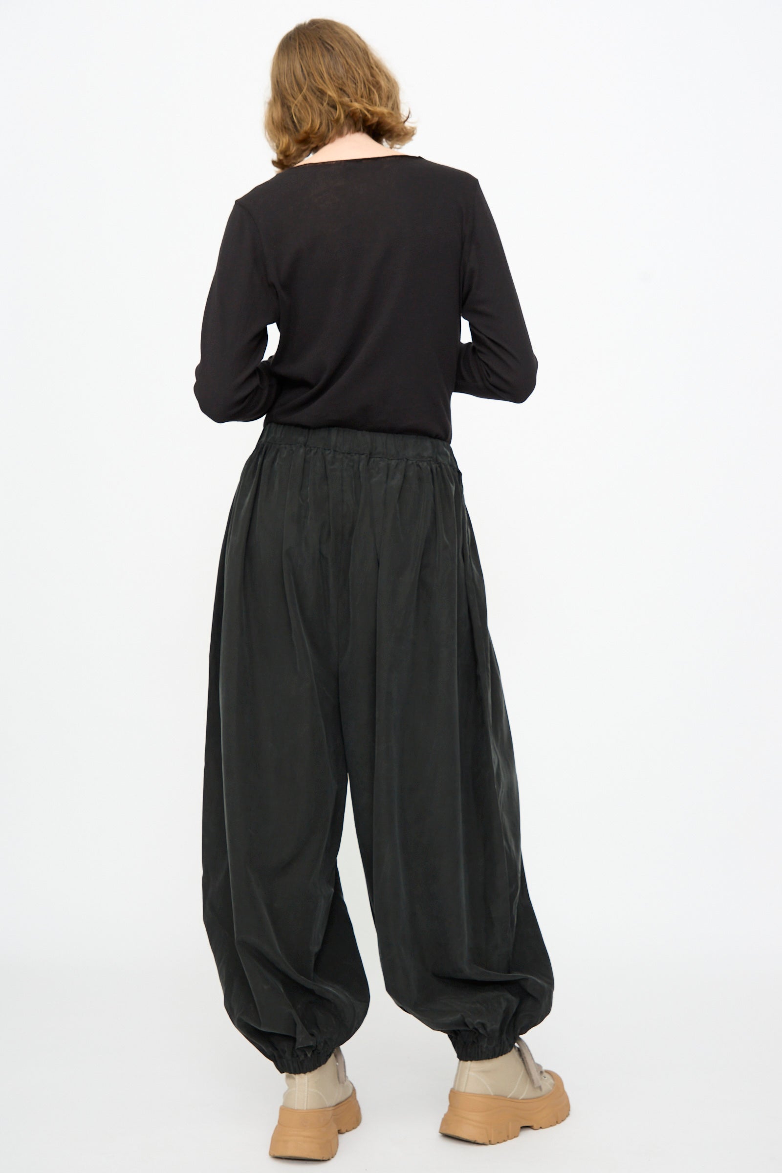 A person stands facing away against a plain white background, wearing Black Crane’s Lantern Pants in Black—a loose silhouette with an elasticated waistband and gathered ankles, crafted from a cotton cupro blend—paired with a black long-sleeve shirt and beige ankle boots.