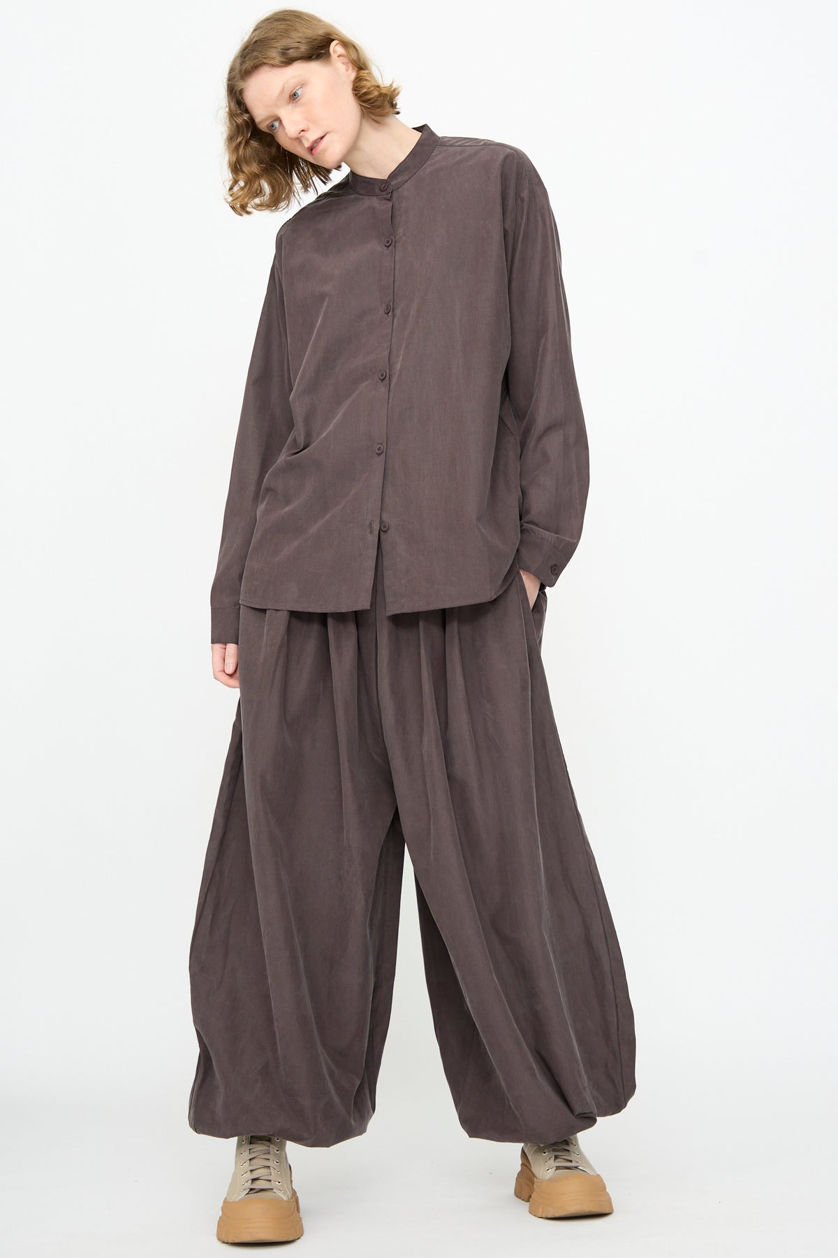 A person wearing Black Crane’s Lantern Pants in Dark Plum—wide, billowy curved leg pants with an elasticated waistband crafted from a soft cotton cupro blend—paired with a loose brown button-up shirt and tan lace-up shoes, standing against a plain white background.