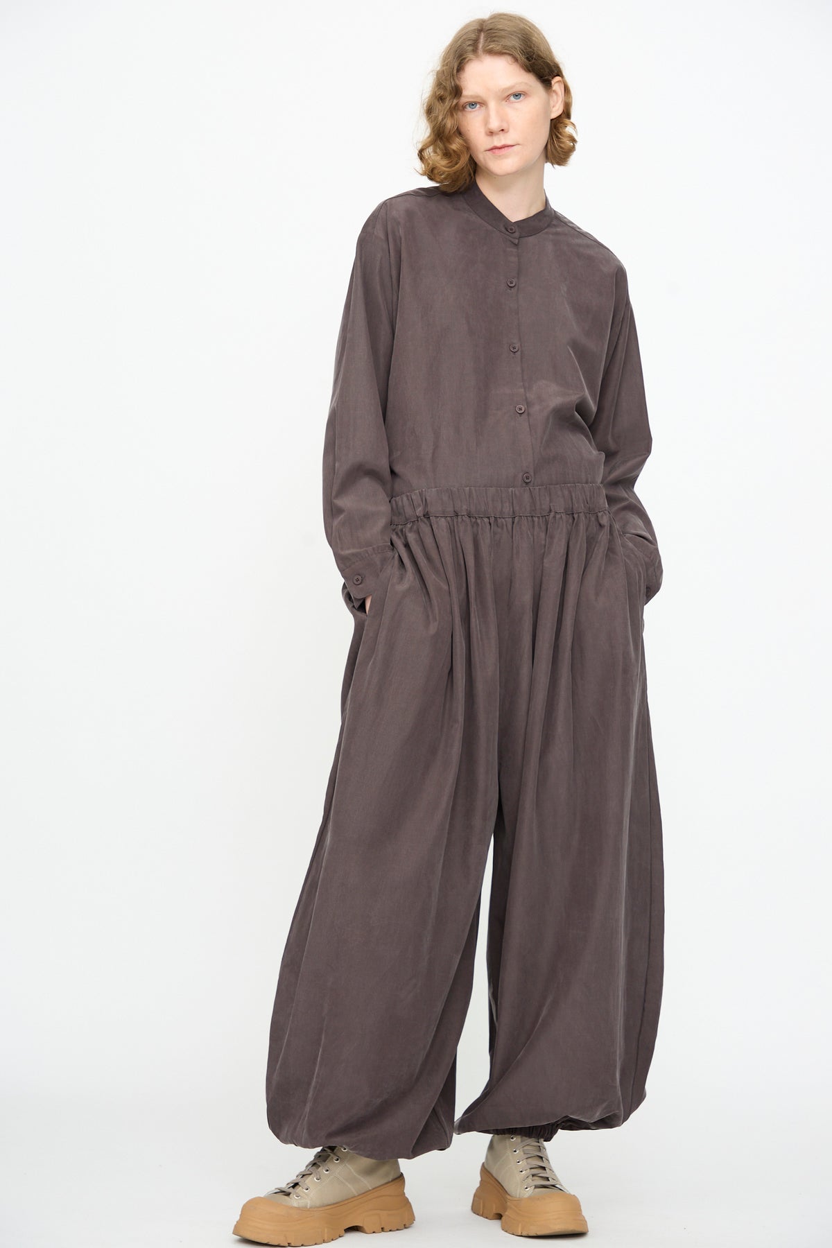 A person stands against a white background, wearing Black Crane’s Lantern Pants in Dark Plum—a loose-fitting, wide, billowy curved-leg style featuring an elasticated waistband—paired with a matching long-sleeve button-up top and tan lace-up shoes.