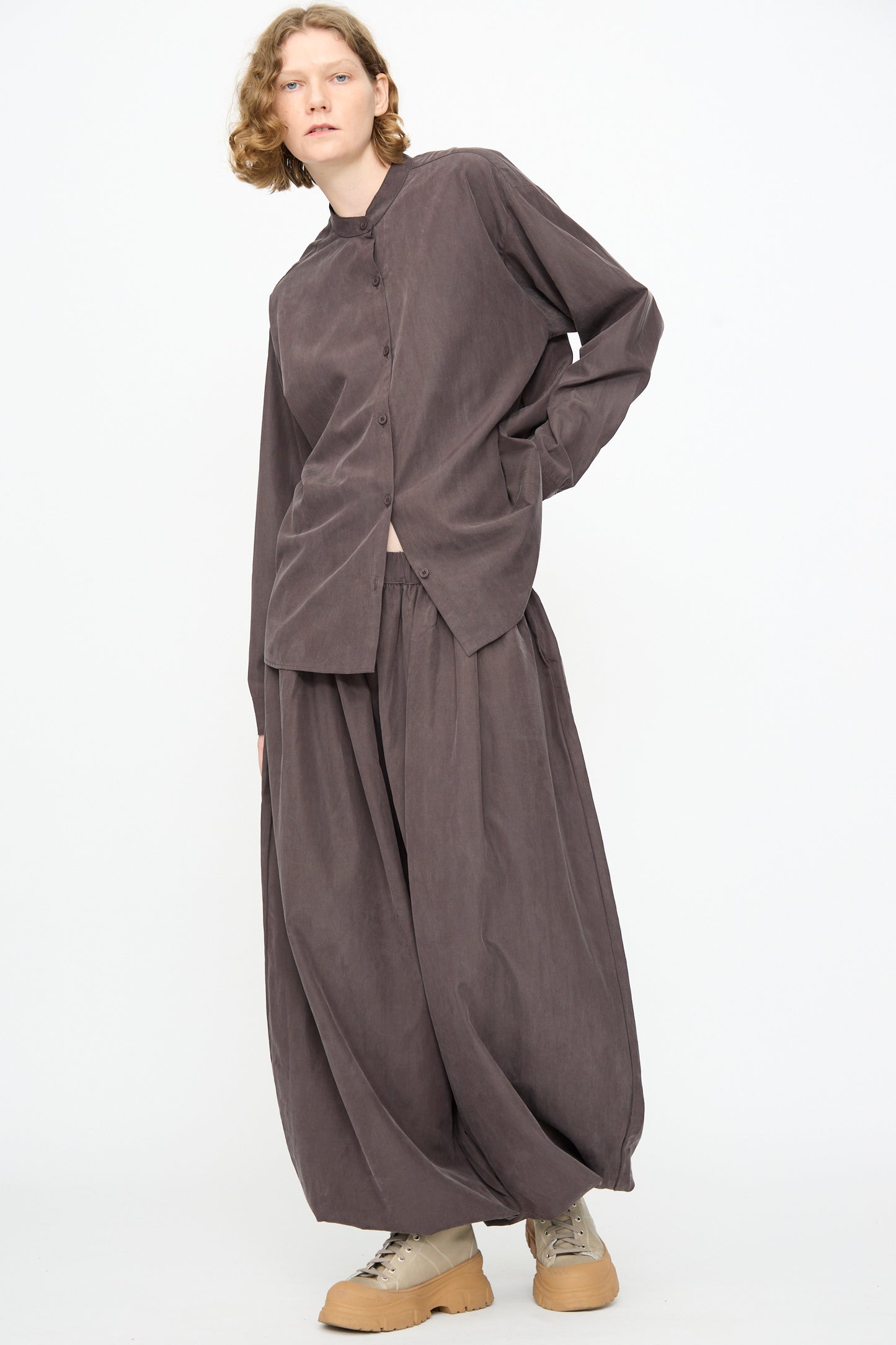 A person is wearing the Black Crane Lantern Pants in Dark Plum, featuring a wide, pleated silhouette with an elasticated waistband and curved leg design in a soft cotton-cupro blend. They are paired with a loose-fitting brown button-up top and beige shoes, standing against a plain white background.