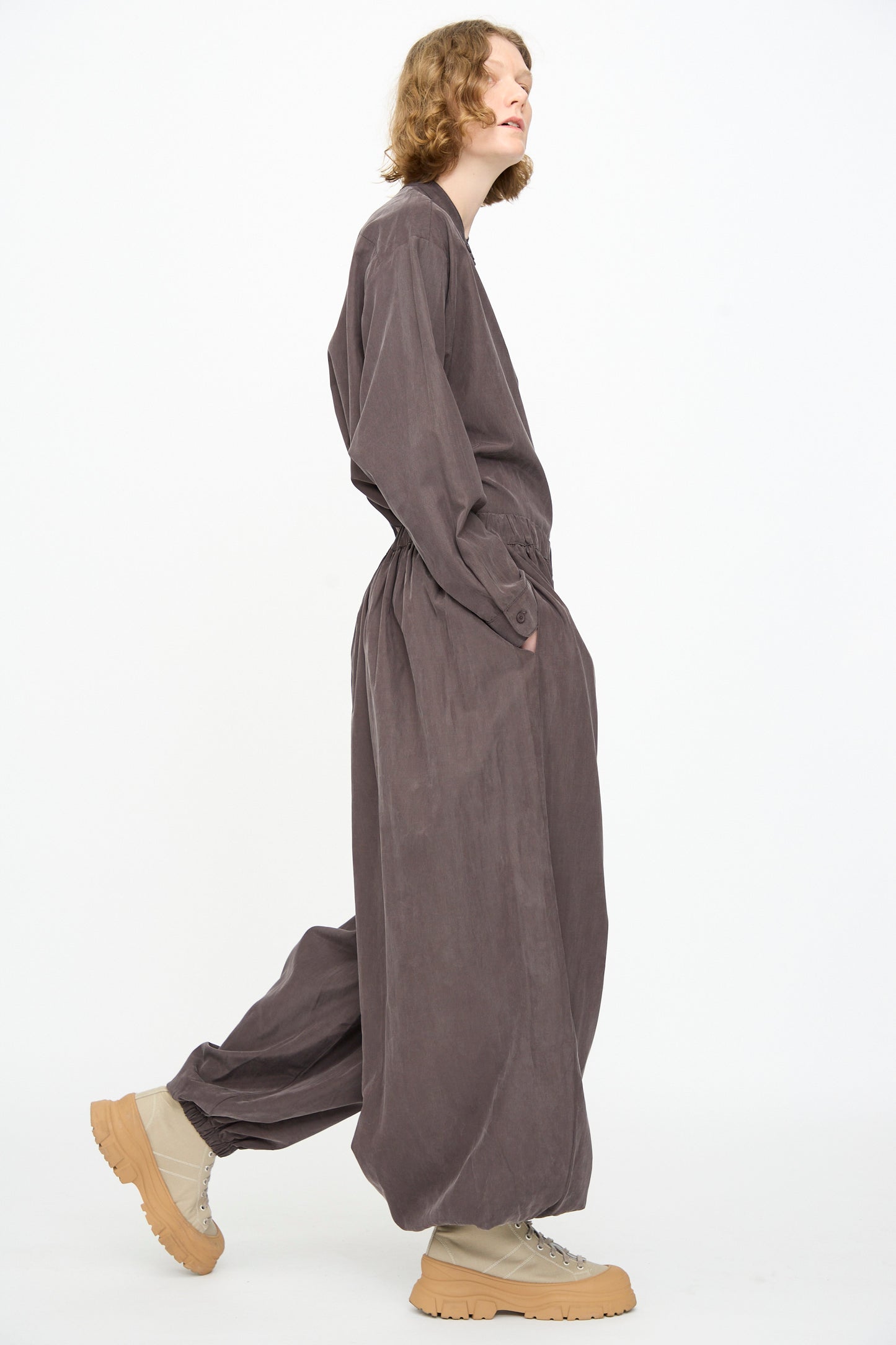 A person stands in profile against a plain white background, wearing the Black Crane Lantern Pants in Dark Plum—a loose-fit style crafted from a soft cotton cupro blend, featuring an elasticated waistband and curved leg silhouette.