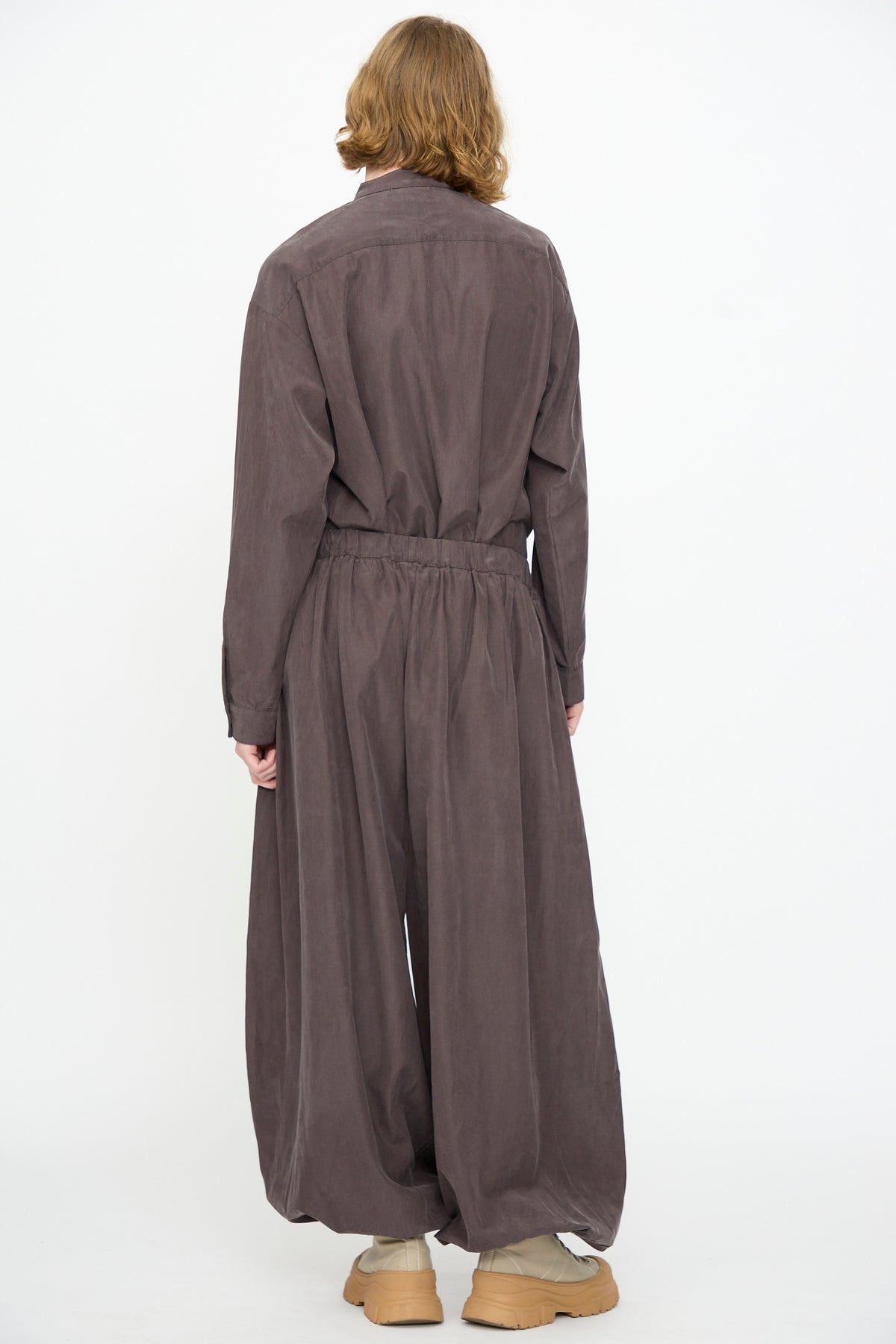 A person stands with their back to the camera, wearing Black Crane’s Lantern Pants in Dark Plum—a loose-fitting, long-sleeved silhouette crafted from a soft cotton cupro blend—featuring a subtle elasticated waistband and beige shoes, set against a plain white background.