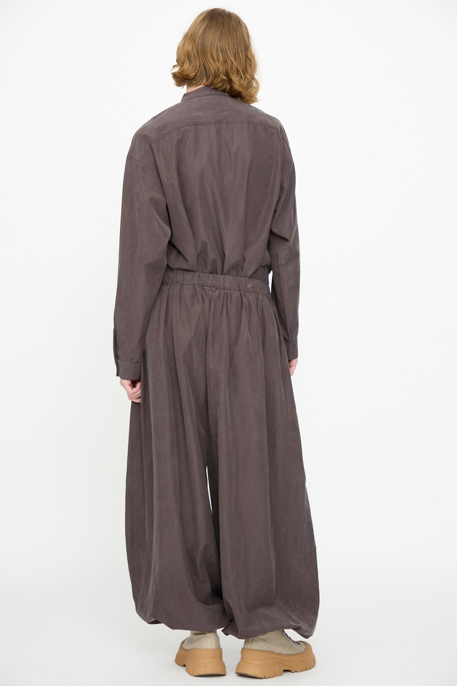 A person stands with their back to the camera, wearing Black Crane’s Lantern Pants in Dark Plum—a loose-fitting, long-sleeved silhouette crafted from a soft cotton cupro blend—featuring a subtle elasticated waistband and beige shoes, set against a plain white background.