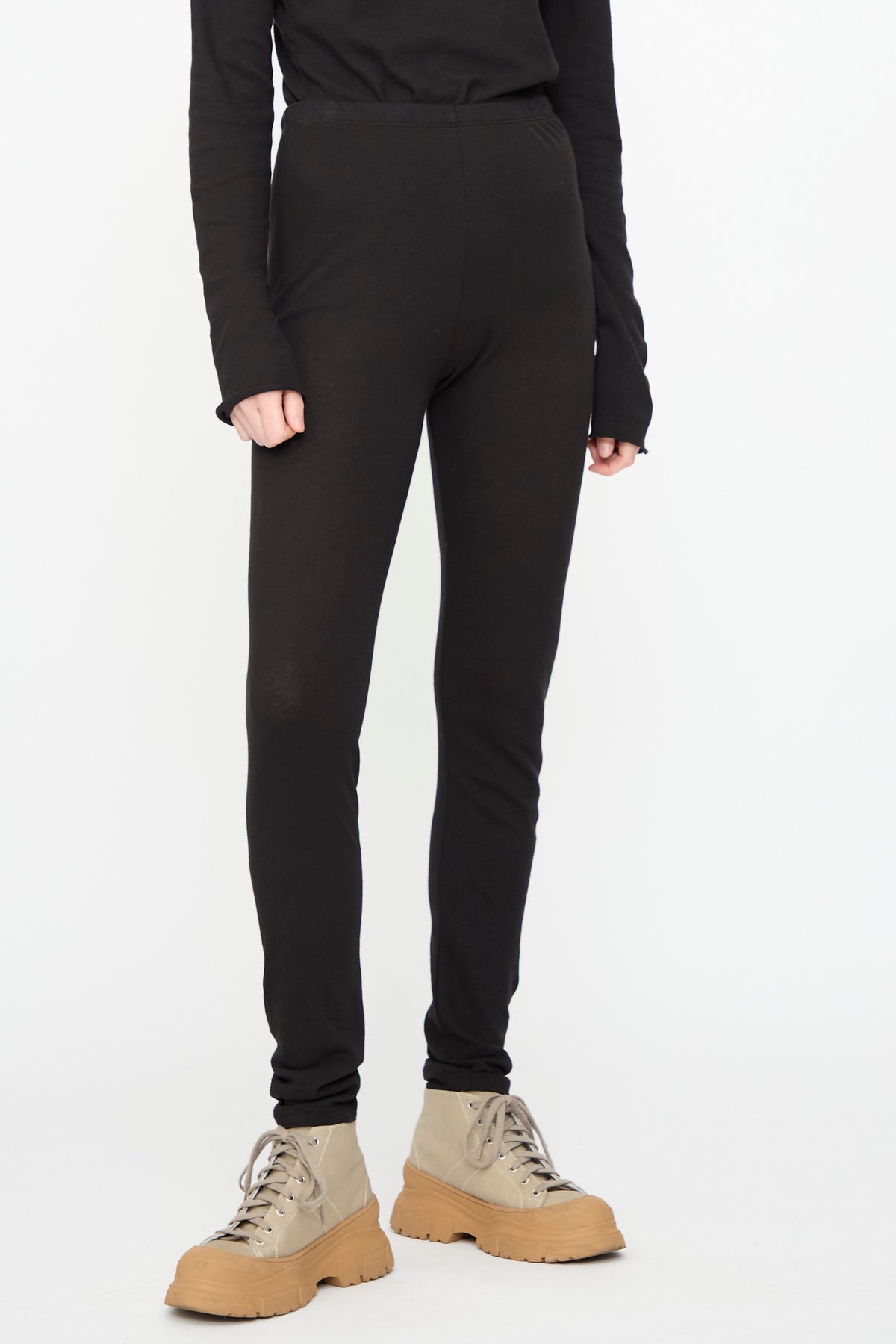 A person stands against a white background, wearing a black long-sleeve top, Black Crane Leggings in Black (made in the USA), and beige lace-up boots. The upper body is partially out of frame.
