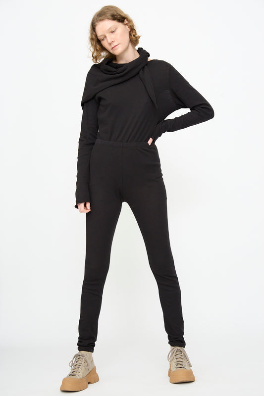 A person in Black Crane’s Leggings in Black stands against a plain white background, wearing a black long-sleeve jumpsuit and tan lace-up shoes, with one hand on their hip and the other relaxed—effortlessly showcasing the sleek silhouette of these slim fit leggings.