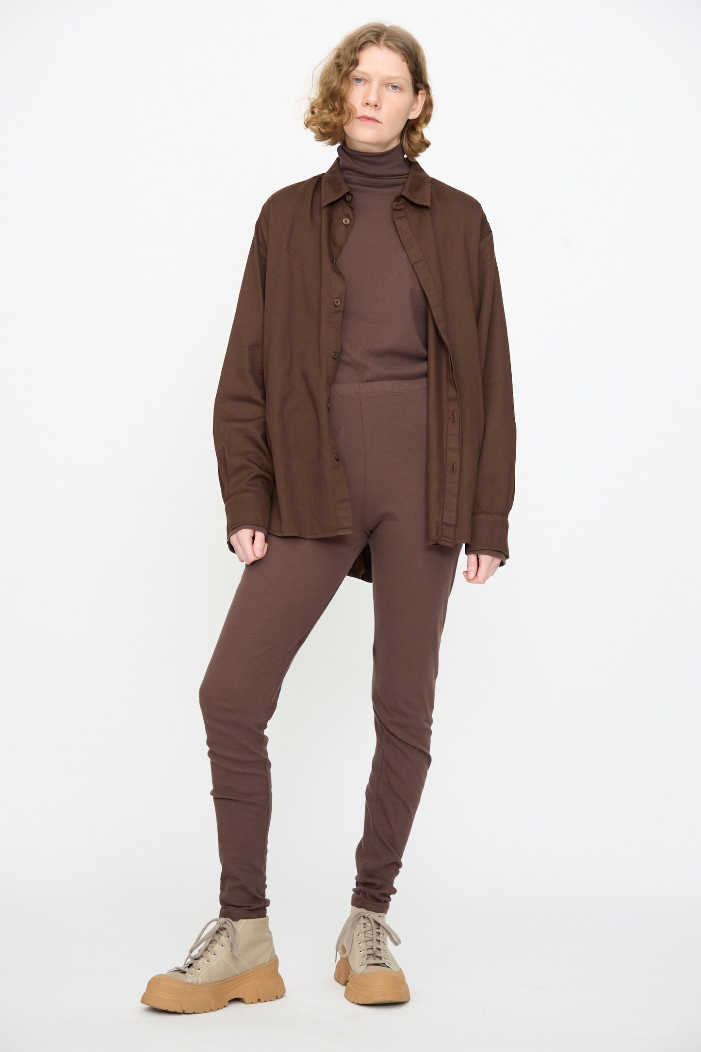 A person stands against a white background, wearing Black Crane’s Leggings in Dark Plum paired with a matching turtleneck and shirt, styled with beige lace-up boots.
