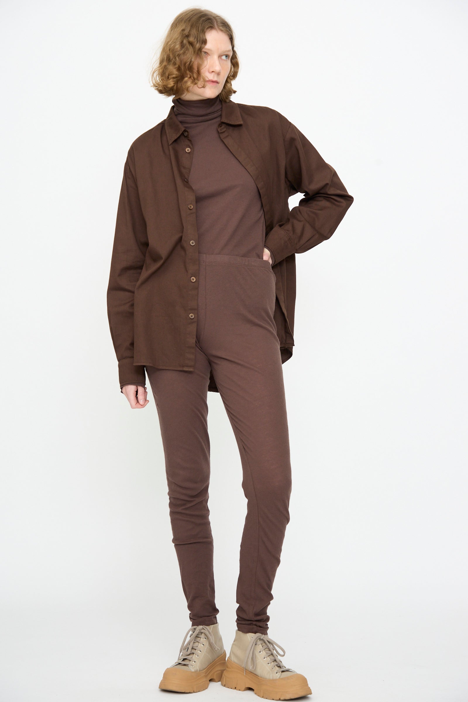 A person stands against a white background, wearing a brown button-up shirt over a brown turtleneck jumpsuit, paired with Black Crane Leggings in Dark Plum and beige lace-up shoes.