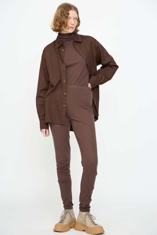 A person stands against a white background, wearing a brown button-up shirt over a brown turtleneck jumpsuit, paired with Black Crane Leggings in Dark Plum and beige lace-up shoes.