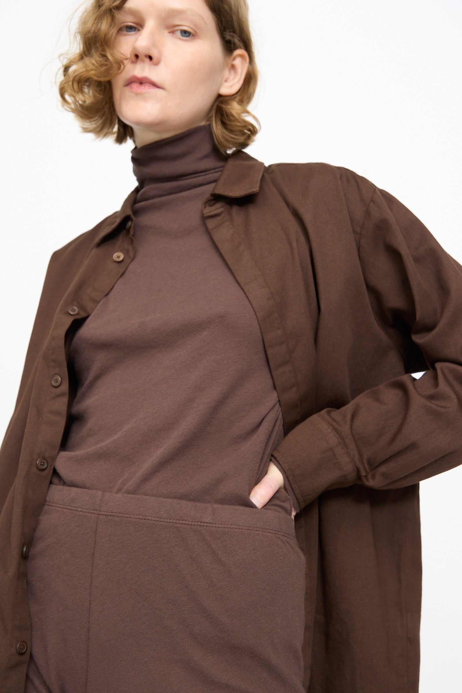 A person wearing a brown button-up shirt over a brown turtleneck and Black Crane Leggings in Dark Plum stands with one hand in their pocket against a plain background.