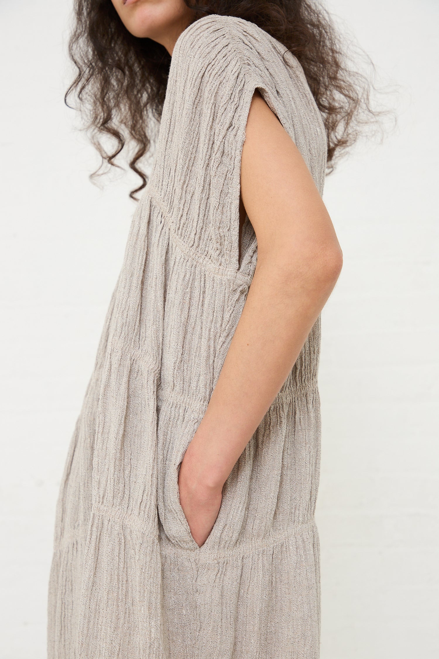 A person in a Linen Accordion Jumpsuit in Ash from Black Crane with their hand in the pocket, standing against a white wall.