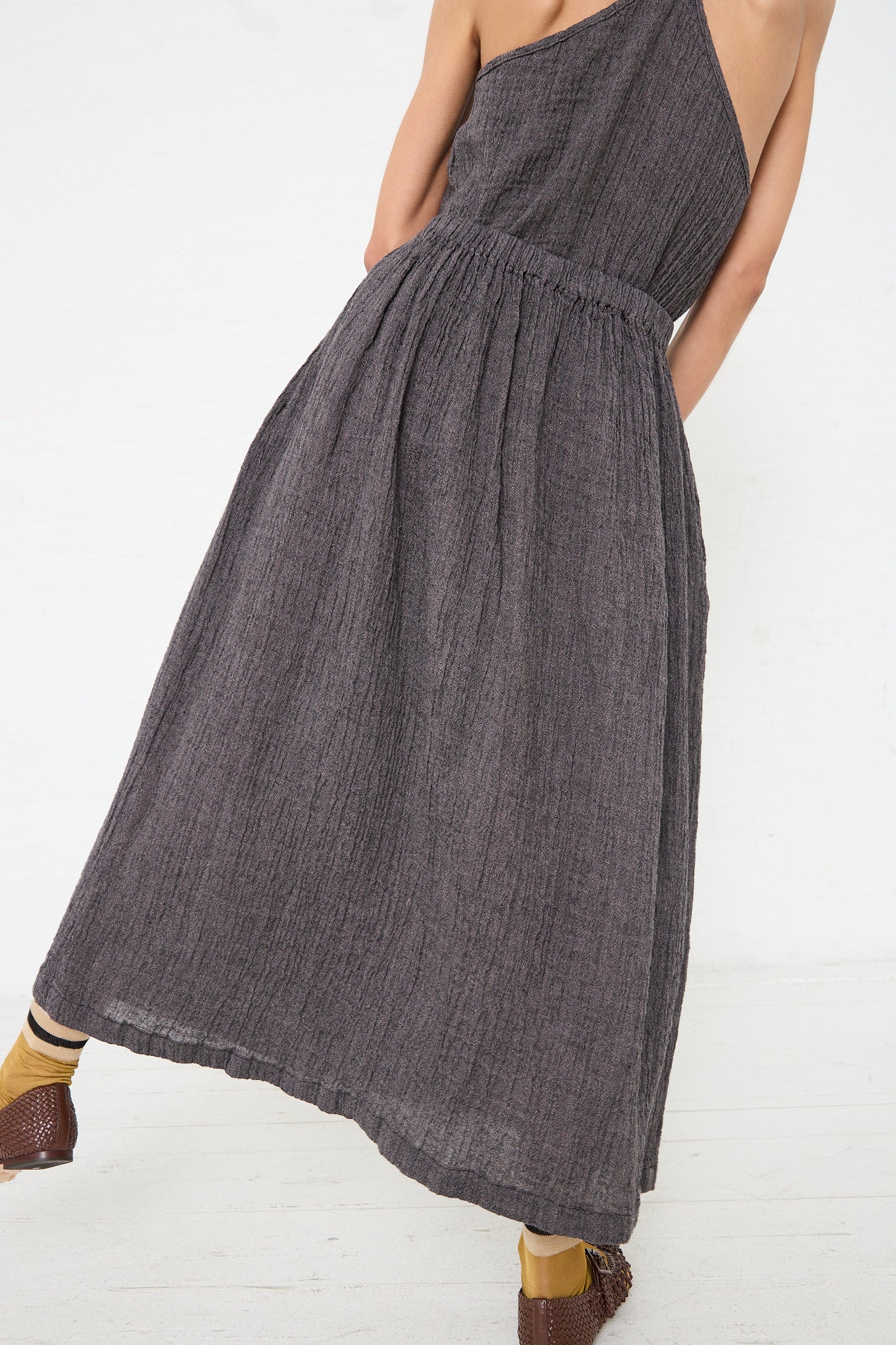 A person wearing a one-shoulder, full-length grey dress with a Linen Classic Skirt in Grey Navy by Black Crane and brown woven platform sandals by Black Crane.