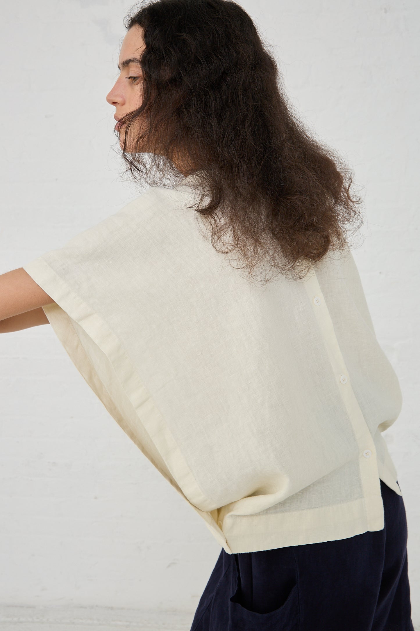 A woman with long dark hair is viewed from the side while adjusting a Black Crane Linen Origami Top in Cream with a boxy fit on her torso. The background is a plain white wall.