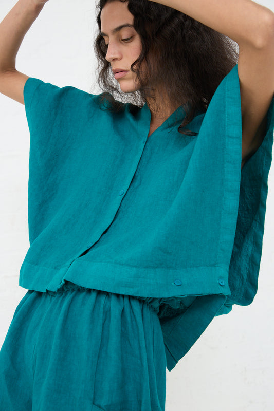 A woman in a Linen Origami Top in Peacock from Black Crane with a boxy fit and pants posing with her arms raised and her head tilted to the side against a white background.