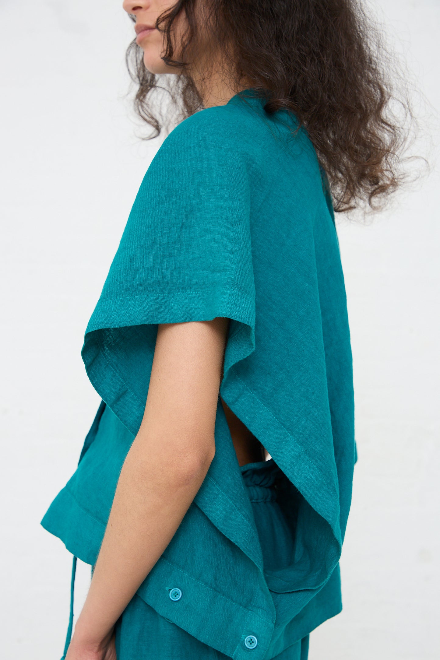 A woman standing in profile, wearing a Linen Origami Top in Peacock by Black Crane, with a boxy fit, the back partly open and button details, against a white background.