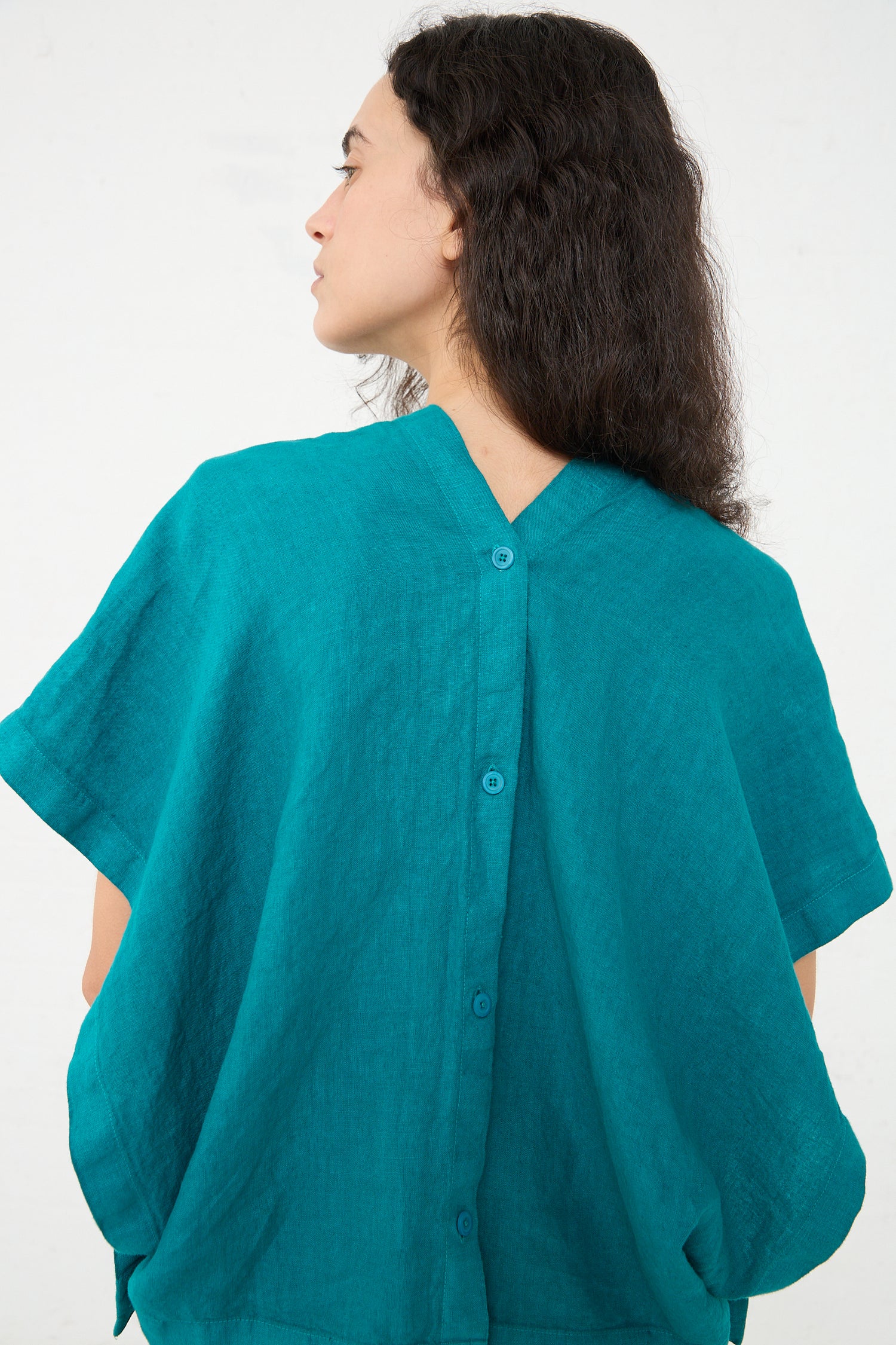 A woman wearing a Linen Origami Top in Peacock by Black Crane, with button details on the back, standing in profile against a white background.