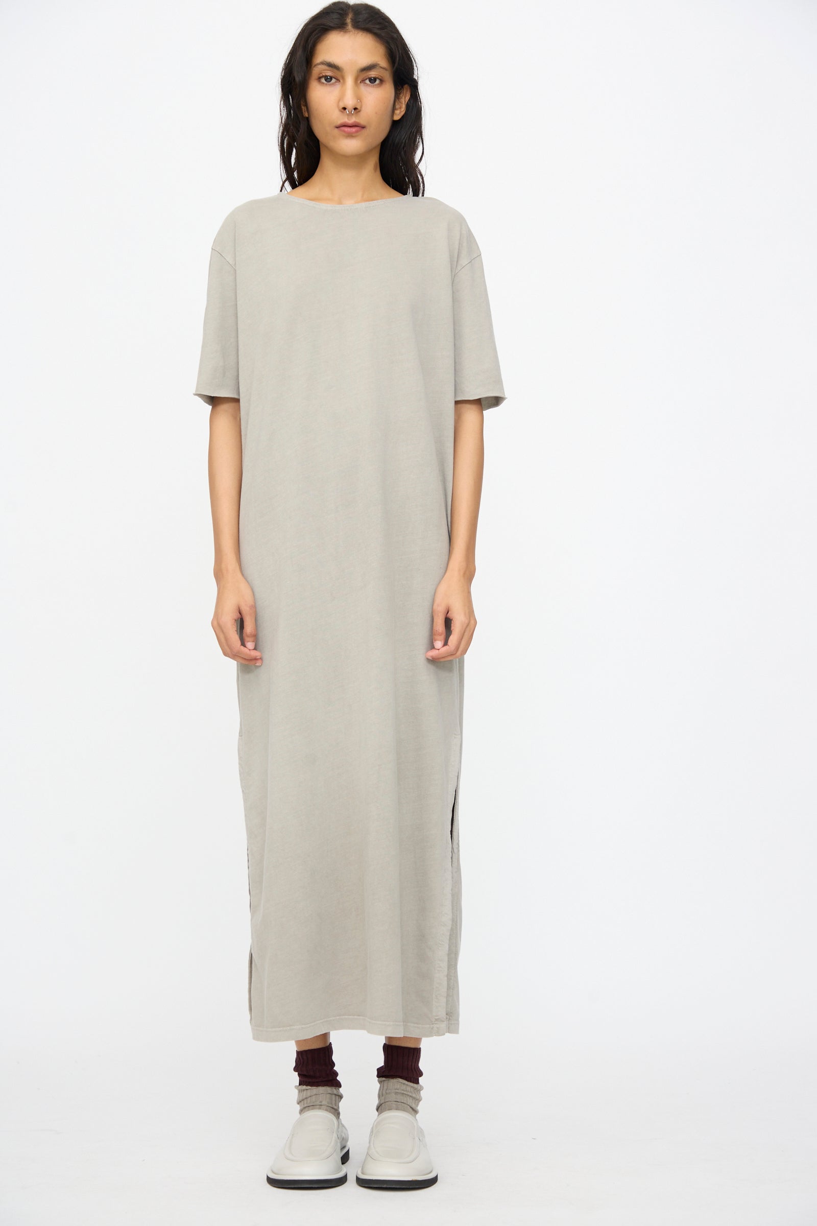A person stands against a plain background wearing the Black Crane Long Tee Dress in Grey—an ankle-length, loose-fitting organic cotton dress with short sleeves and side slits—paired with brown socks and beige slip-on shoes.