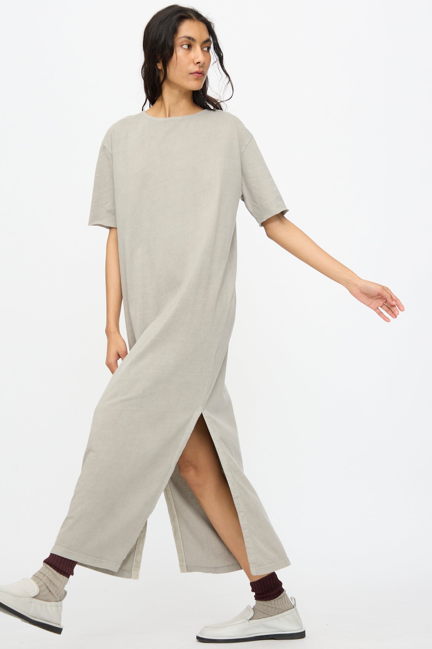 Someone wearing the Black Crane Long Tee Dress in Grey—an organic cotton, loose fit maxi with side slits—paired with dark socks and white sneakers, walking against a plain white background.
