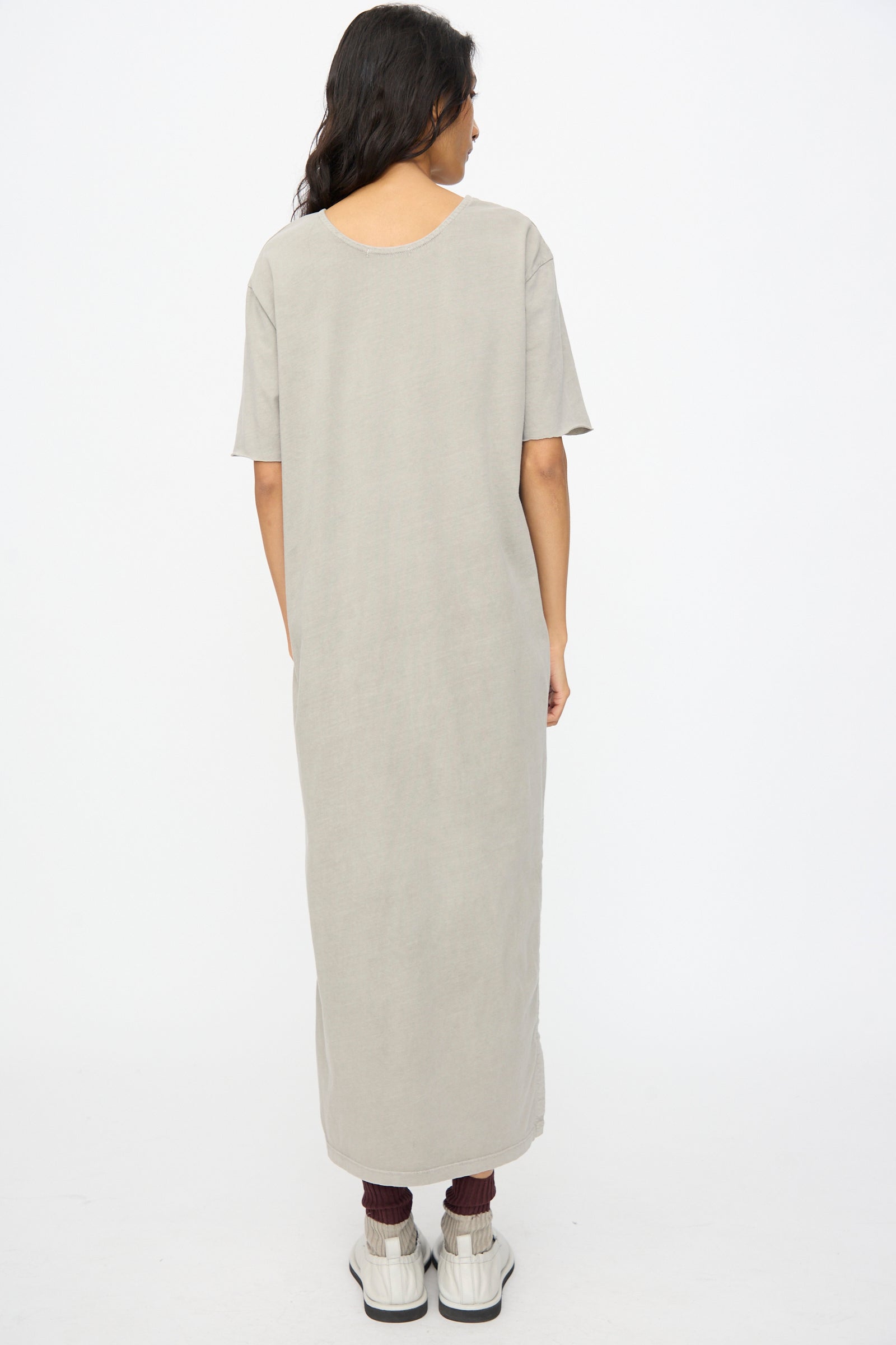 A person stands facing away, wearing the Black Crane Long Tee Dress in Grey—an ankle-length, loose fit organic cotton short-sleeve dress—with white shoes and maroon socks against a plain white background.