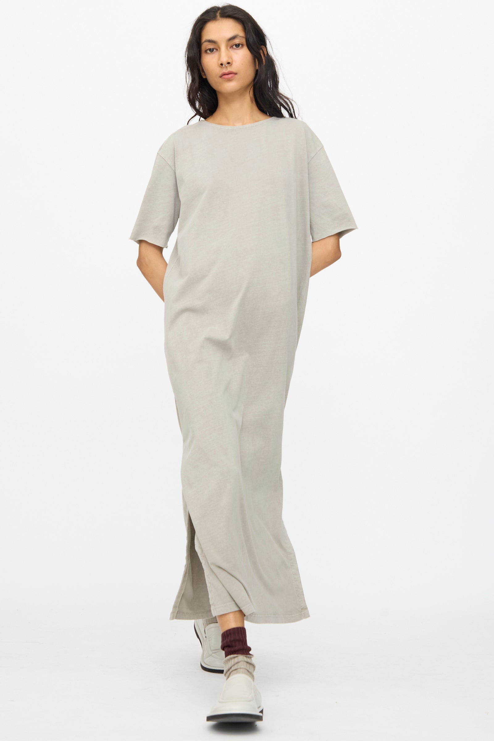 A person wears the Black Crane Long Tee Dress in Grey, a loose fit organic cotton piece, paired with brown socks and white shoes, standing against a plain white background.