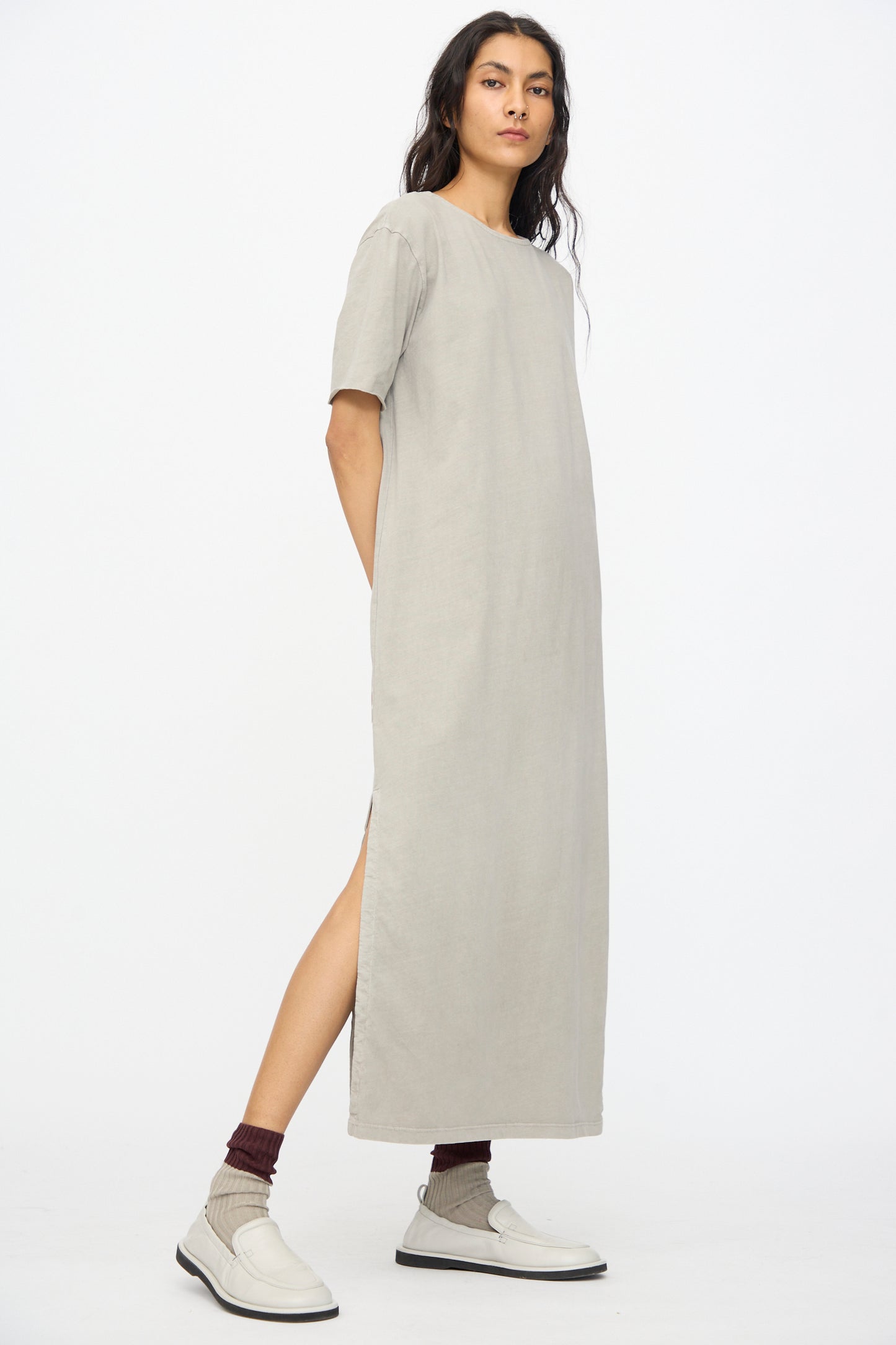 A person wears the Black Crane Long Tee Dress in Grey, a loose fit short-sleeved maxi with side slits, styled with maroon socks and white loafers, standing against a plain white background.