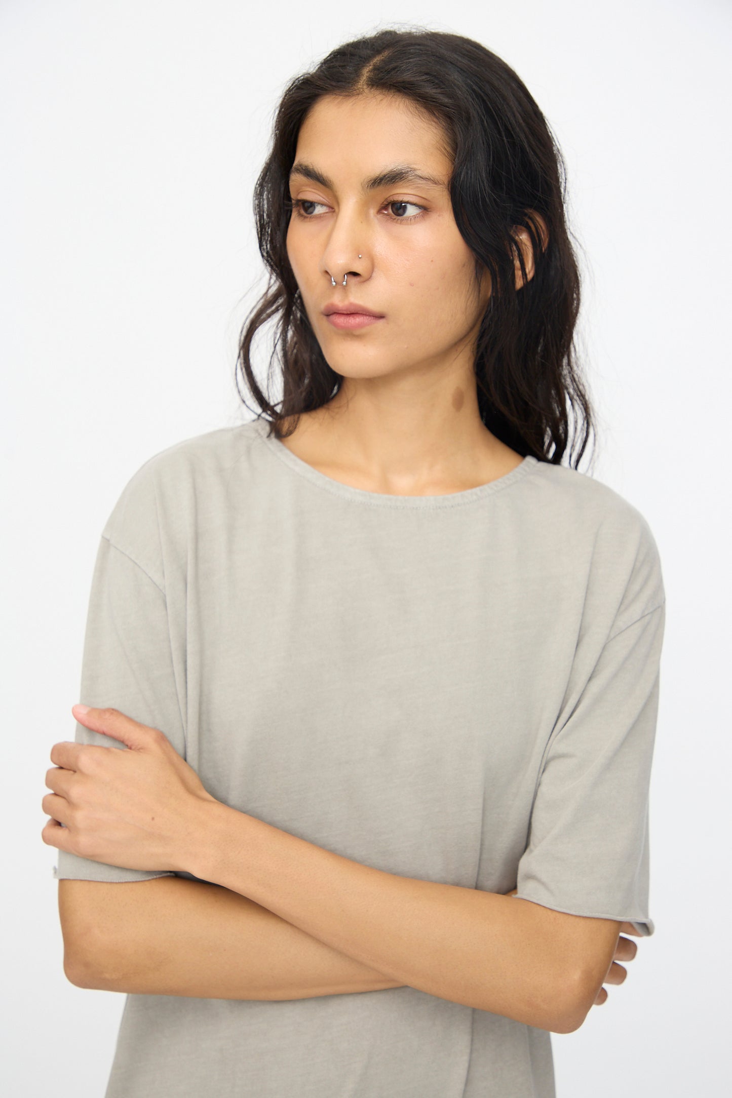 A person with long dark hair and a nose ring wears the Black Crane Long Tee Dress in Grey, standing with arms crossed against a plain white background.