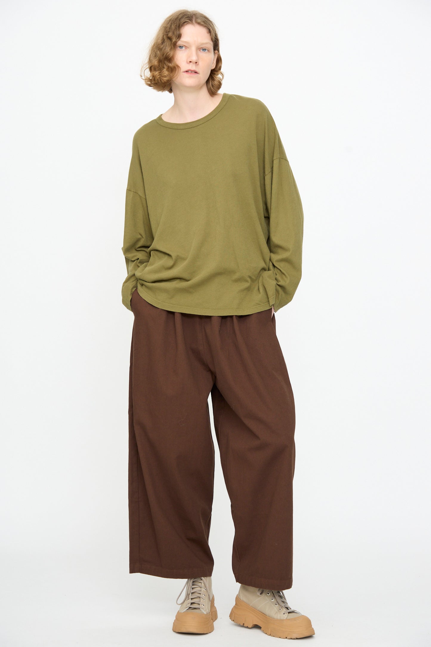 A person stands against a white background wearing the Black Crane Loose Fit Tee in Army—a lightweight, oversized cotton long sleeve shirt—paired with loose brown pants and beige lace-up shoes. Hands are in pockets.