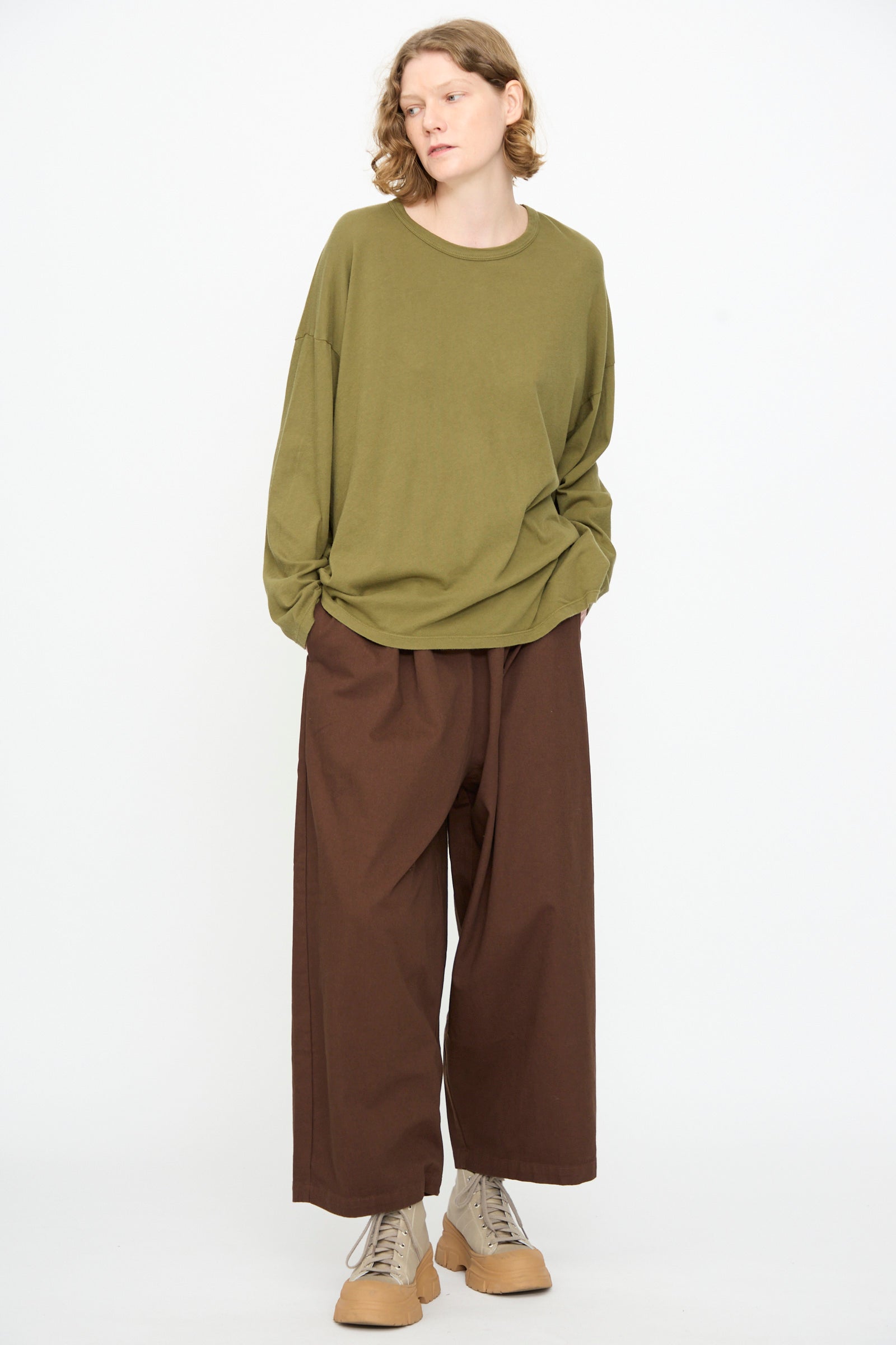 A person wearing the Black Crane Loose Fit Tee in Army—an oversized olive green long sleeve shirt crafted from lightweight cotton—paired with wide brown pants and beige sneakers, stands against a plain white background.