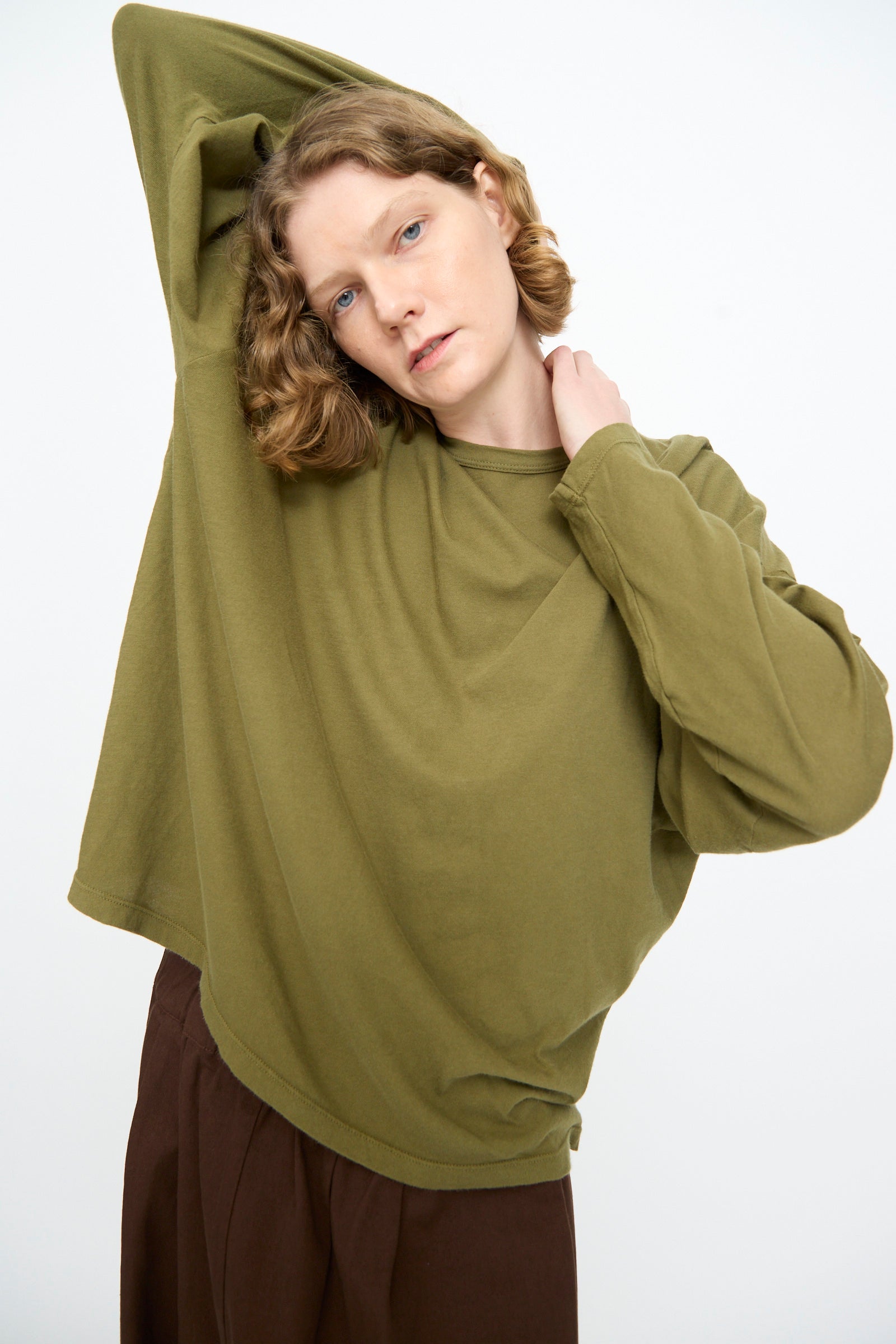 Wearing the Black Crane Loose Fit Tee in Army, a model poses with one arm raised behind their head and pairs the oversized lightweight cotton long sleeve shirt with brown pants against a plain light background.