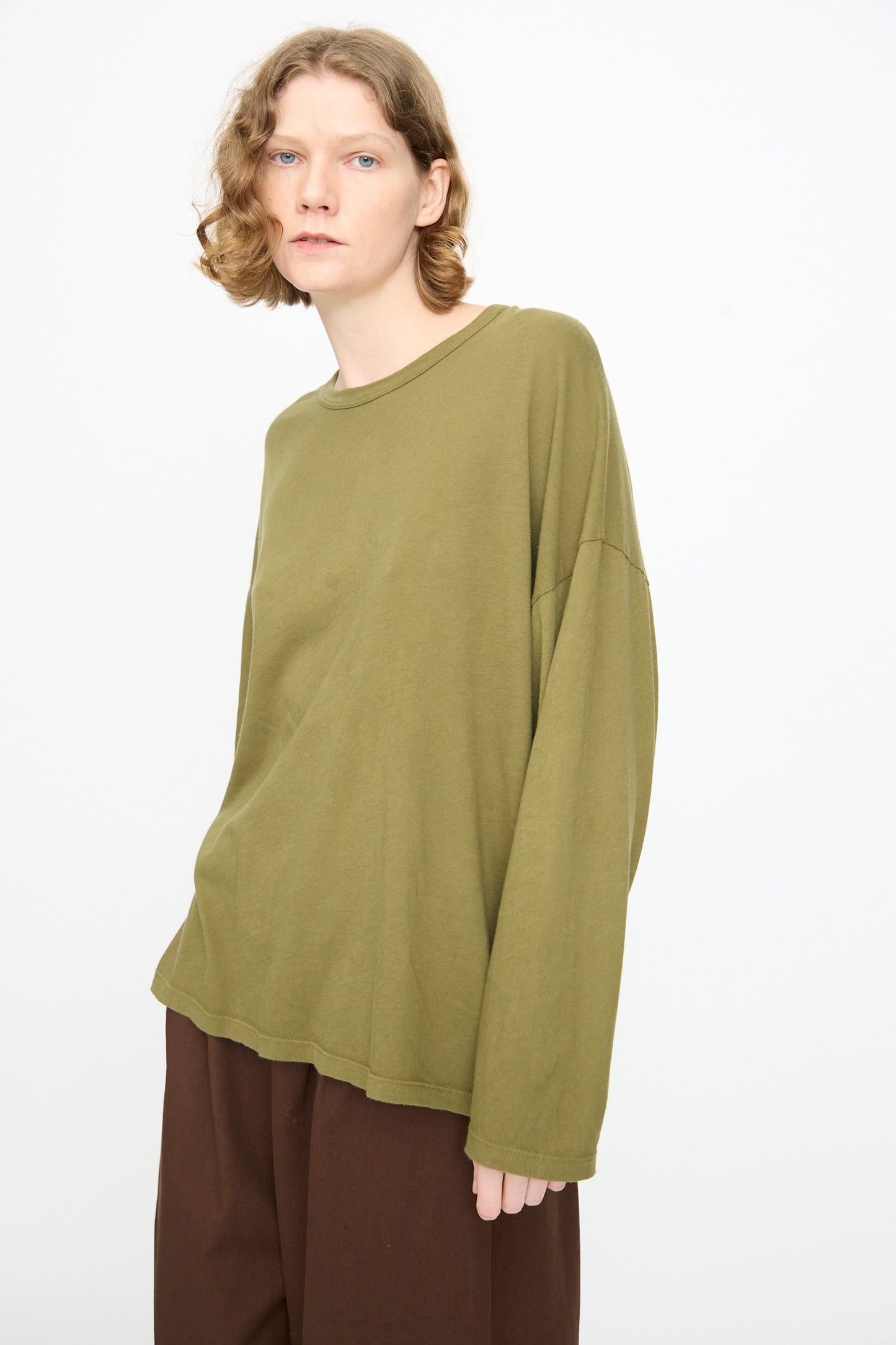 A person with wavy, chin-length hair stands against a plain white background, wearing the Black Crane Loose Fit Tee in Army—a long-sleeve shirt crafted from lightweight cotton—and brown pants.