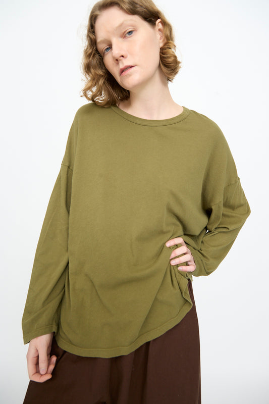 A person with short, wavy hair wears the Black Crane Loose Fit Tee in Army and brown pants, standing against a plain white background.