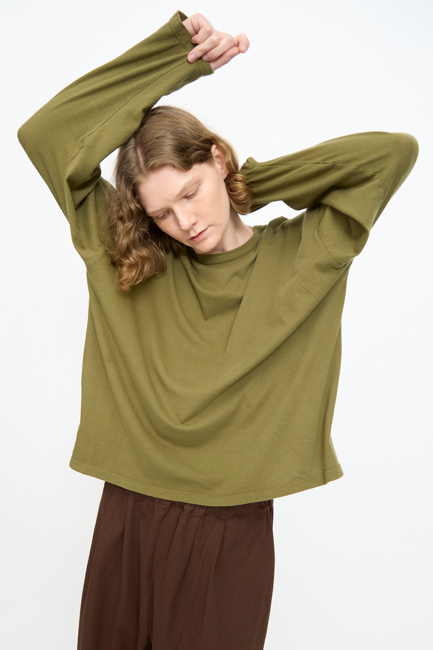 Wearing the Black Crane Loose Fit Tee in Army, a person with brown pants raises one arm and tilts their head downward against a plain white background.