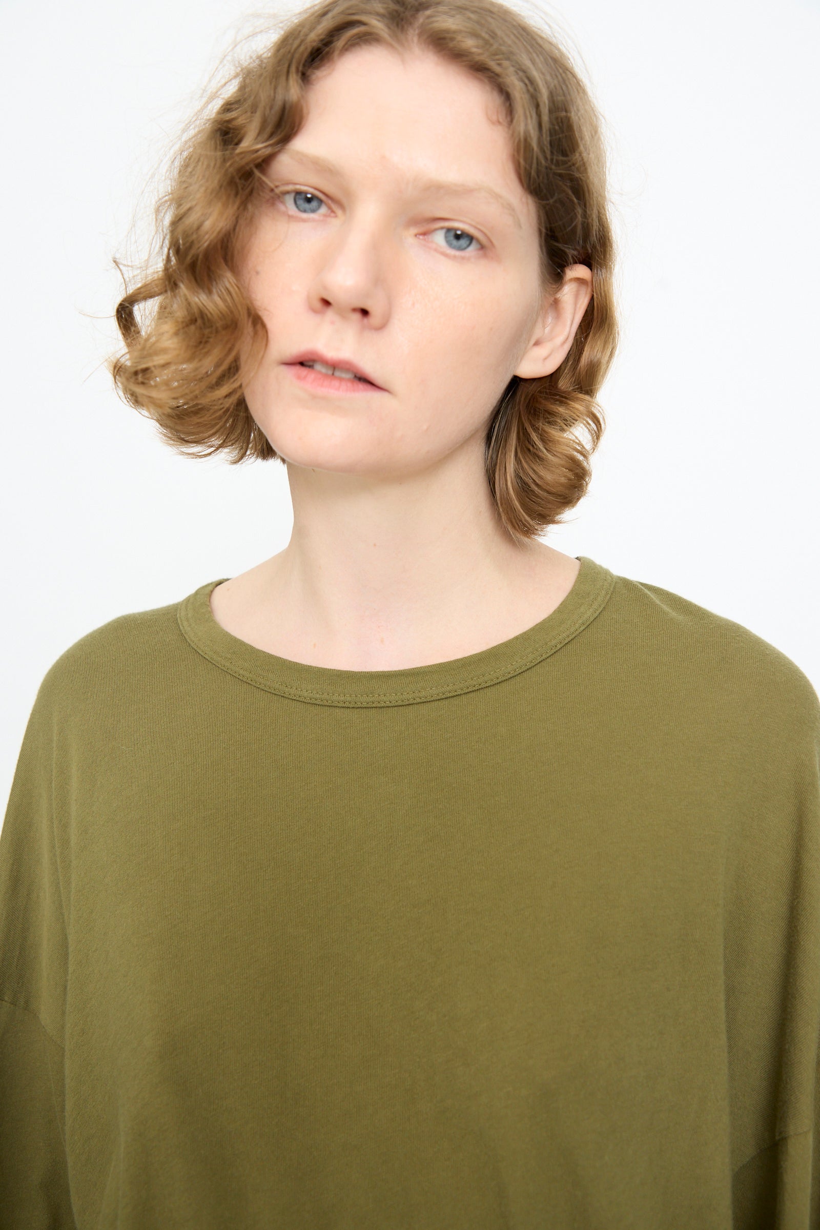 A person with short, wavy light brown hair wears the Black Crane Loose Fit Tee in Army—an olive green, lightweight cotton long sleeve shirt—while standing against a plain white background and looking slightly past the camera.