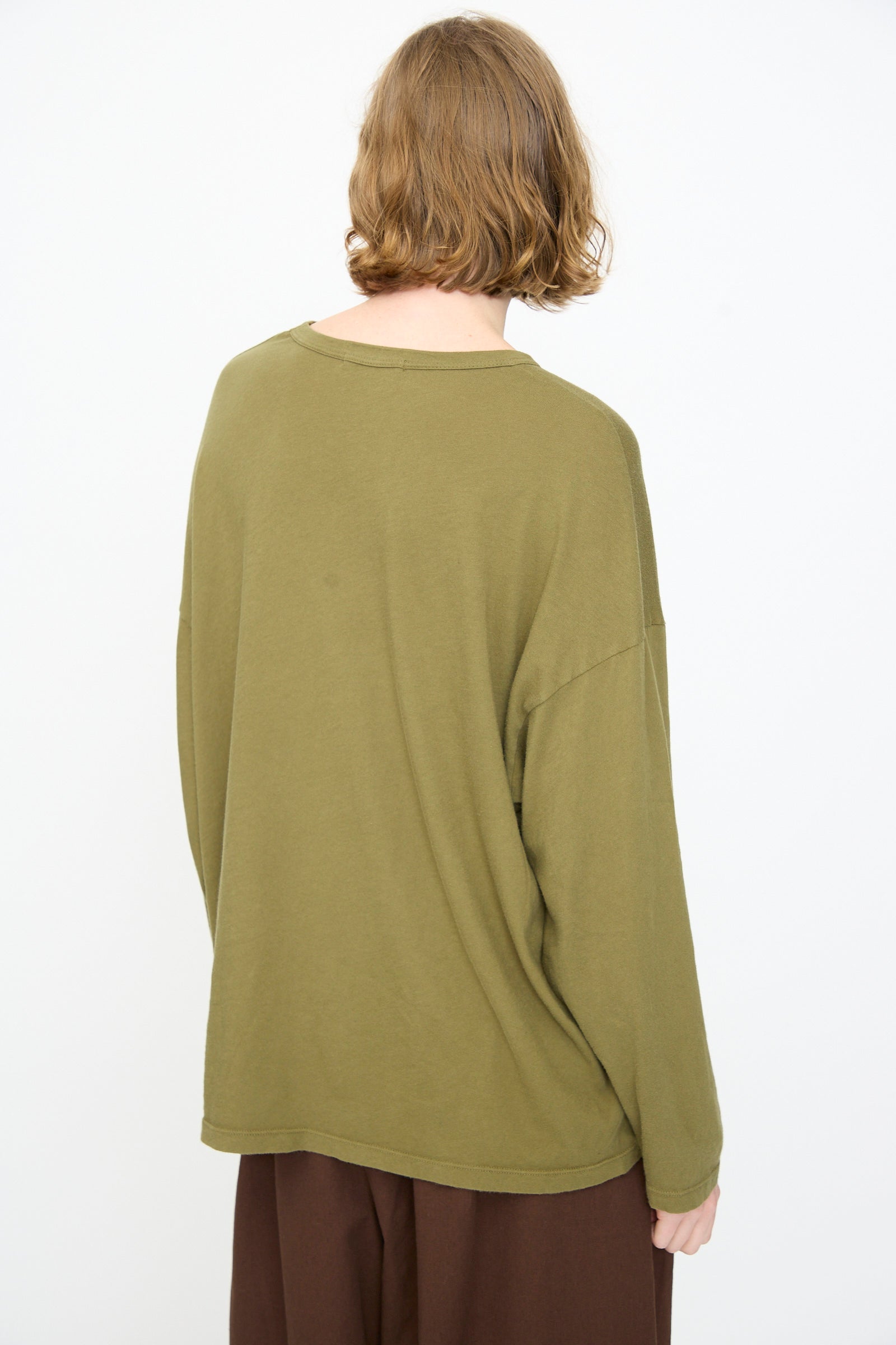A person with short, light brown hair is shown from behind wearing the Black Crane Loose Fit Tee in Army—a lightweight cotton, olive green long sleeve shirt—paired with brown pants, against a plain white background.