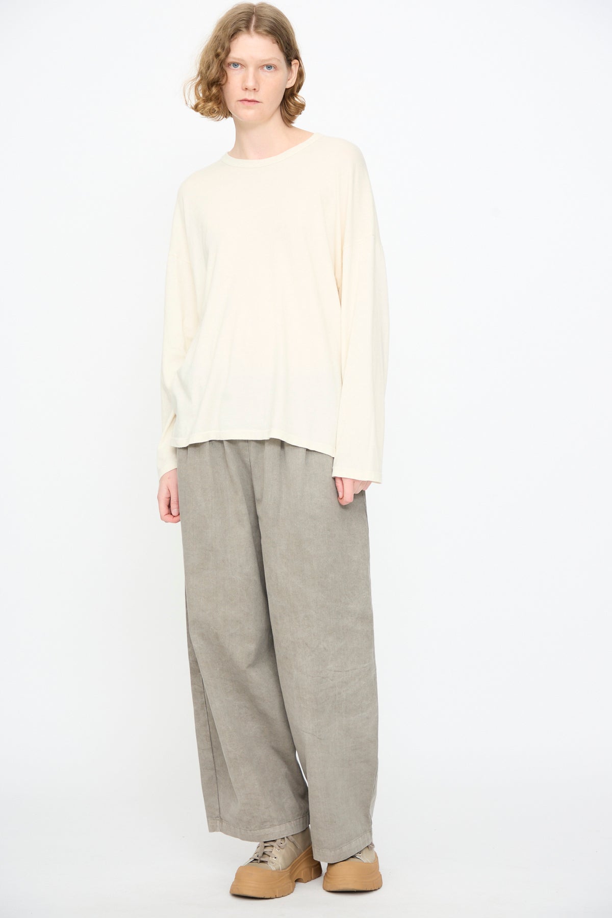 A person stands against a white background, wearing the Black Crane Loose Fit Tee in white, paired with wide gray pants and tan shoes.
