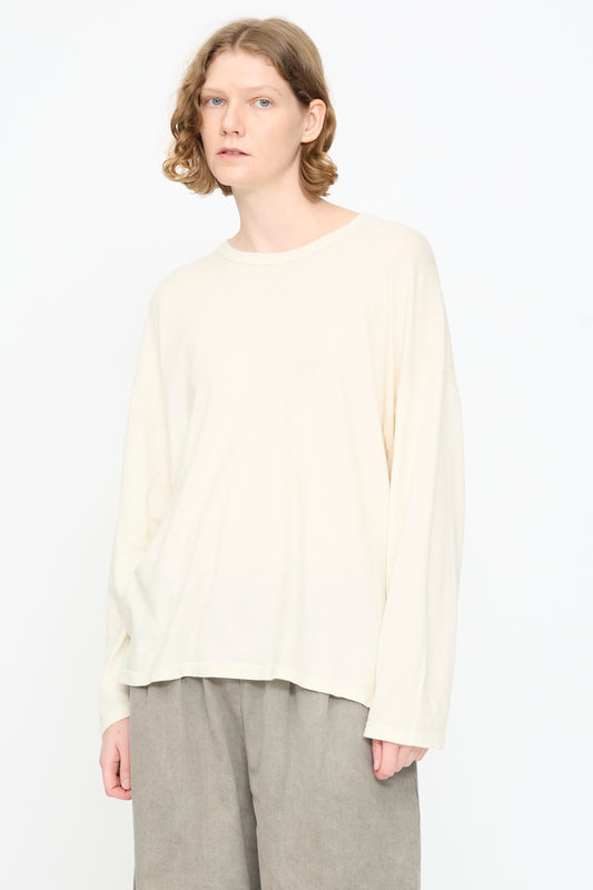 A person with shoulder-length wavy hair is wearing the Black Crane Loose Fit Tee in White and gray pants, standing against a plain white background.