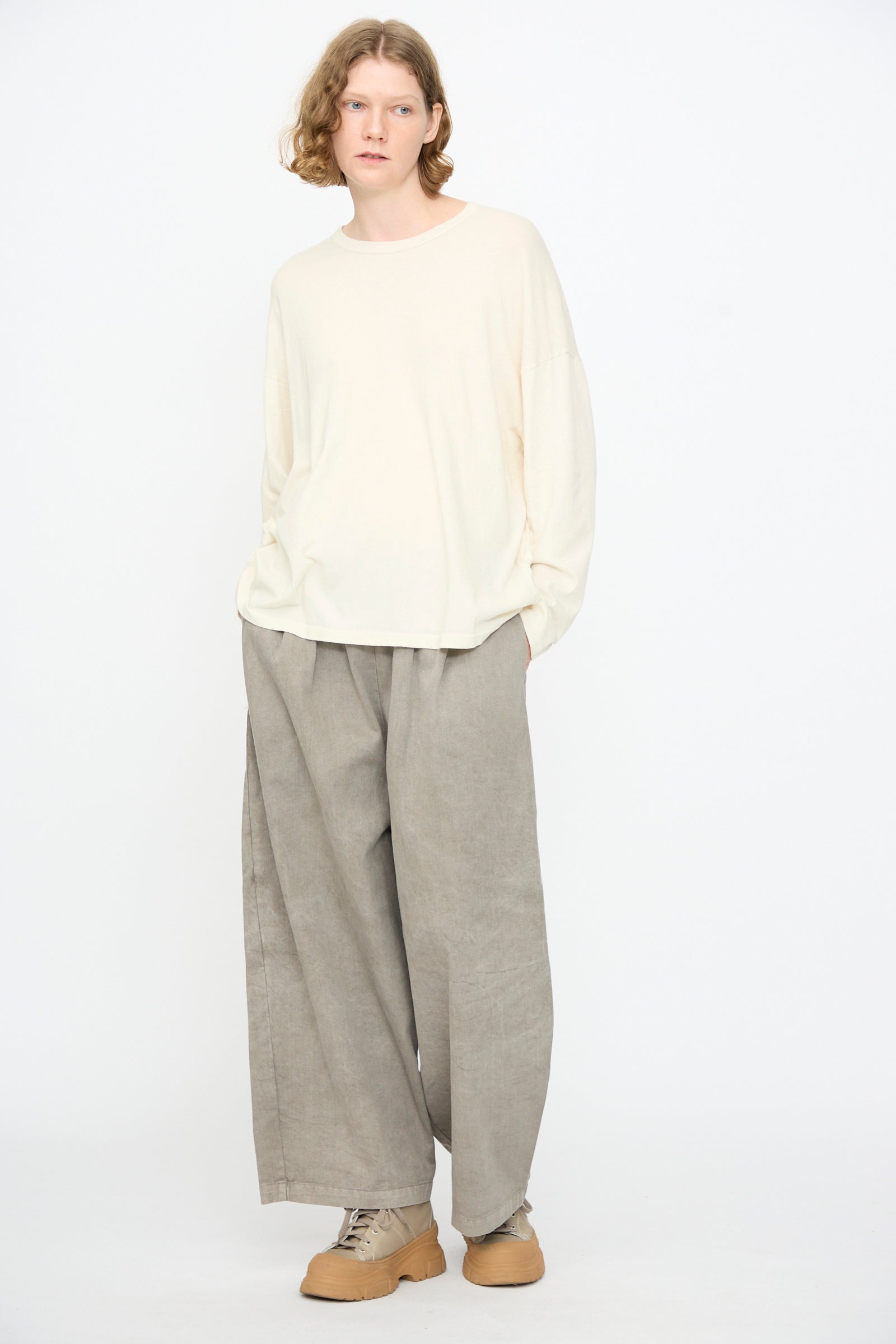 A person wearing the Black Crane Loose Fit Tee in White, made from cotton in the USA, pairs it with wide grey pants and tan shoes, standing against a plain white background with hands in pockets.
