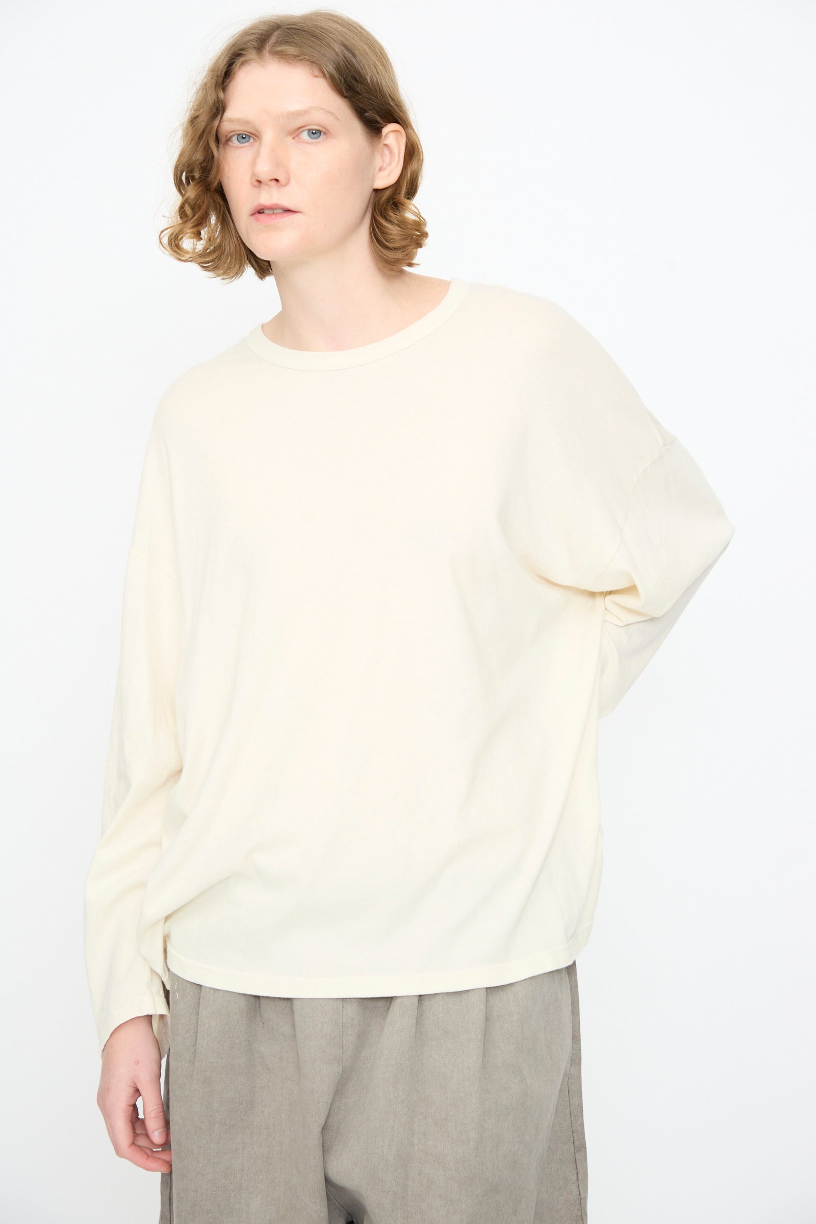A person with shoulder-length light brown hair wears the Black Crane Loose Fit Tee in White, paired with light gray pants, standing against a plain white background.