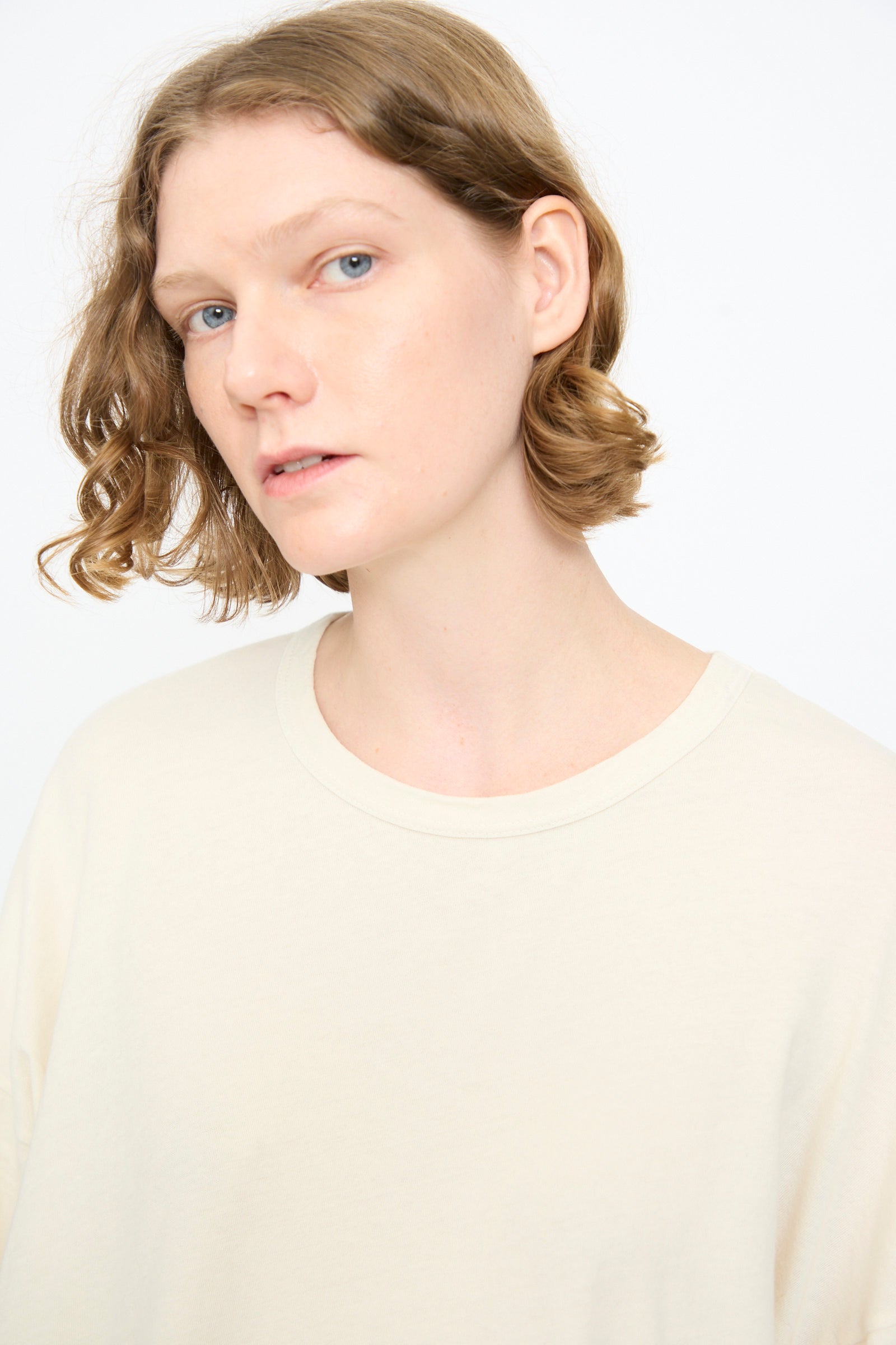 A person with short, wavy, light brown hair and fair skin is wearing the Black Crane Loose Fit Tee in White and looking slightly to the side against a white background.