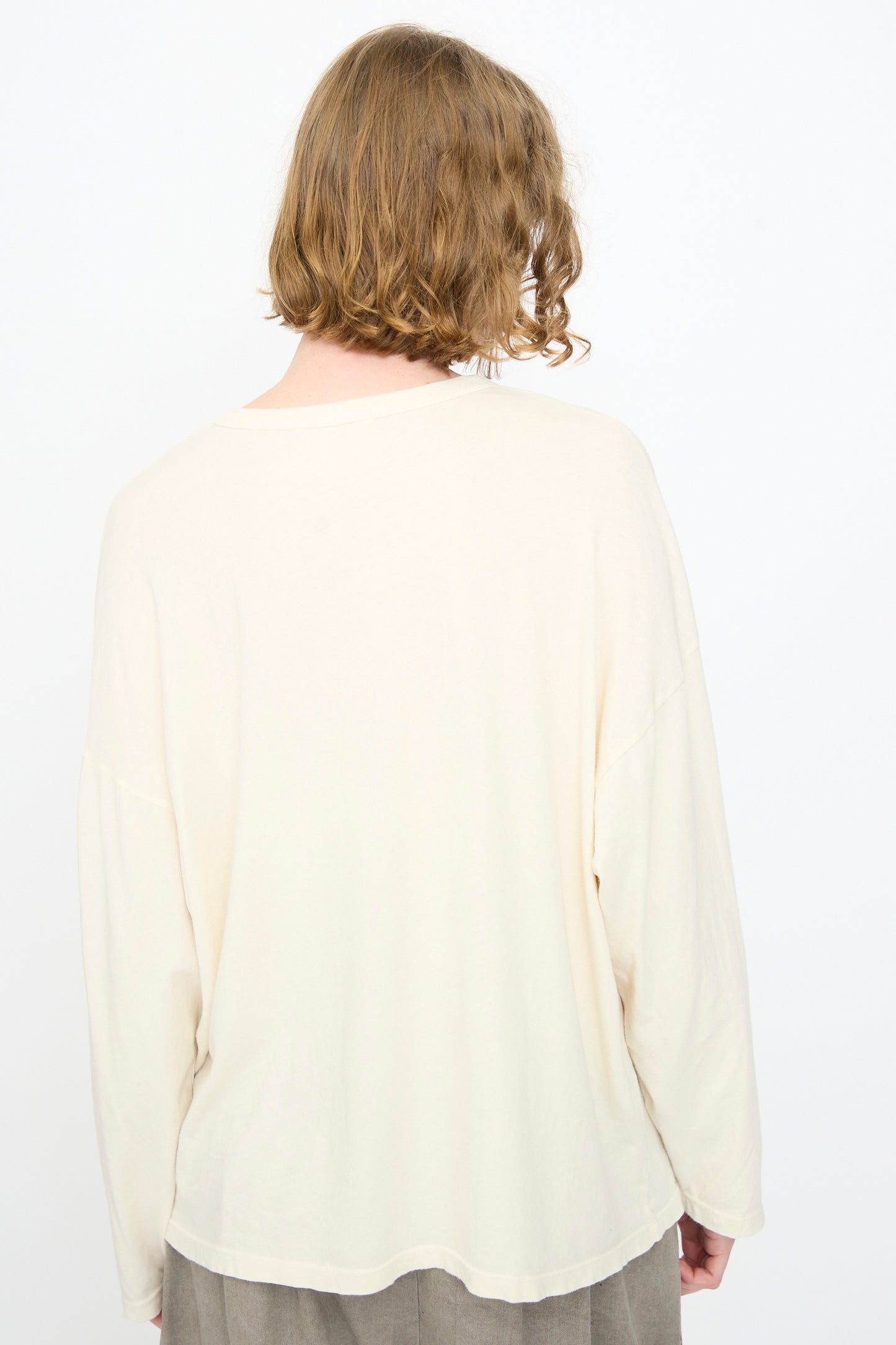 A person with shoulder-length wavy hair, seen from behind against a plain white background, is wearing the Black Crane Loose Fit Tee in White—an oversized cotton long sleeve shirt made in the USA.