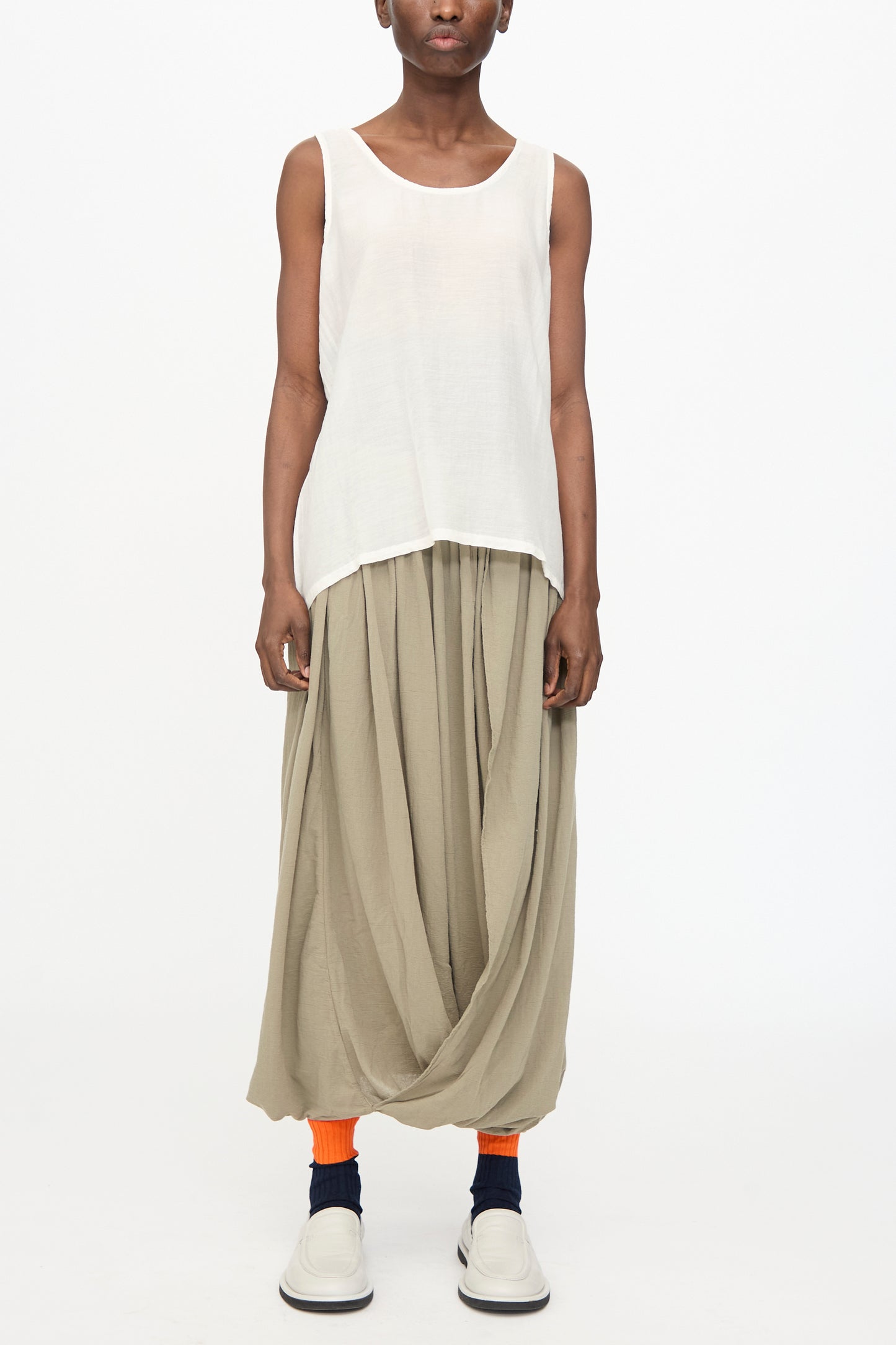 A person wears the Black Crane Loose Tank Top in White, loose beige harem pants, orange socks, and white slip-on shoes, standing against a plain white background.