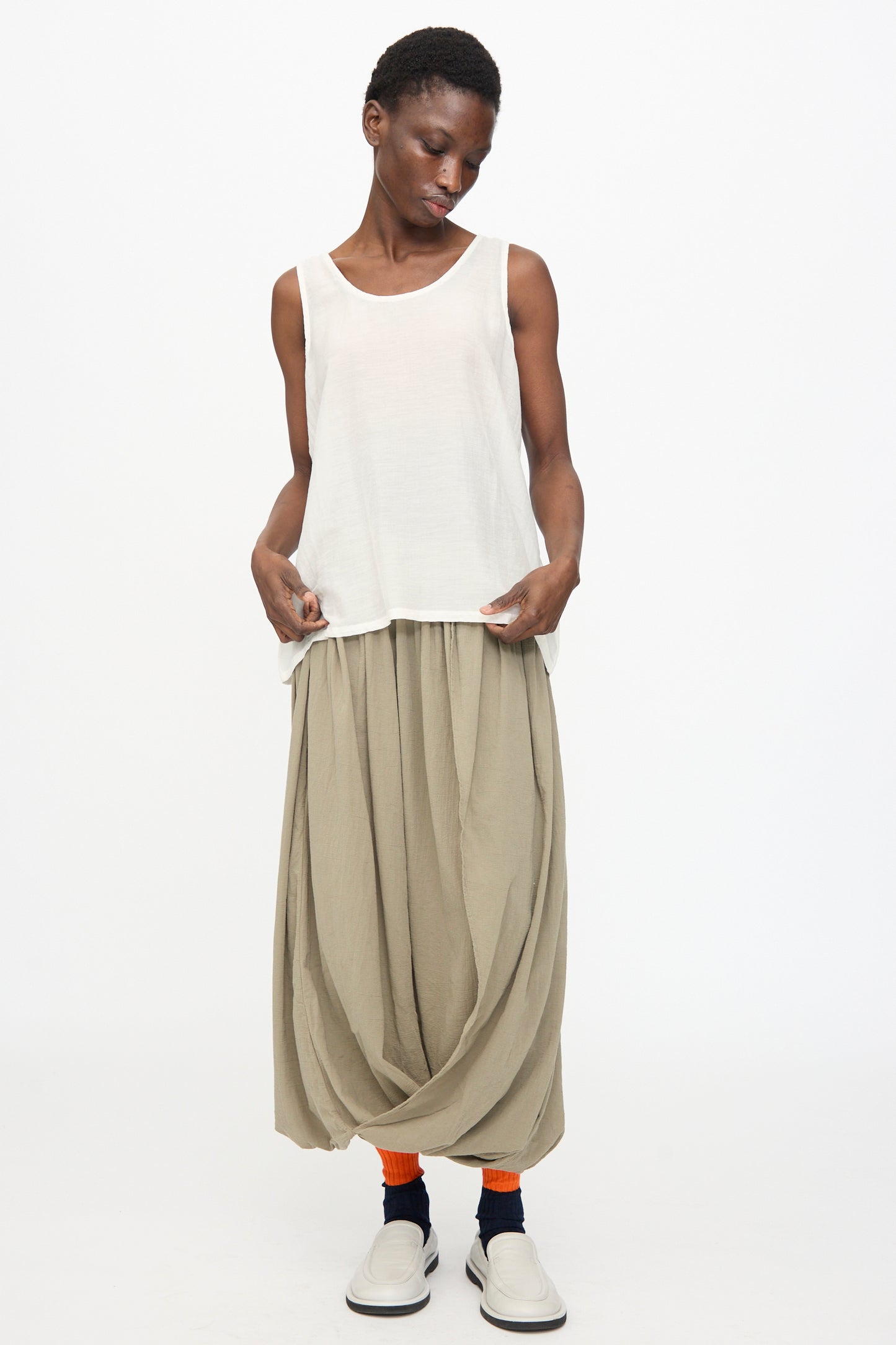A person wears the Black Crane Loose Tank Top in White, a loose beige draped skirt, orange socks, black ankle bands, and white slip-on shoes, standing against a plain white background.