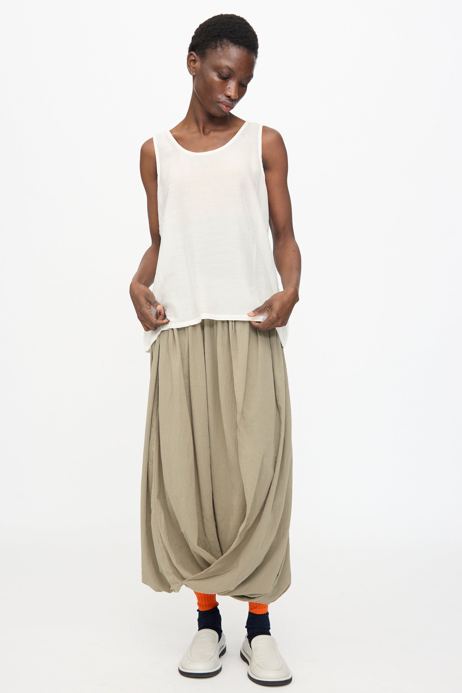 A person wears the Black Crane Loose Tank Top in White, a loose beige draped skirt, orange socks, black ankle bands, and white slip-on shoes, standing against a plain white background.