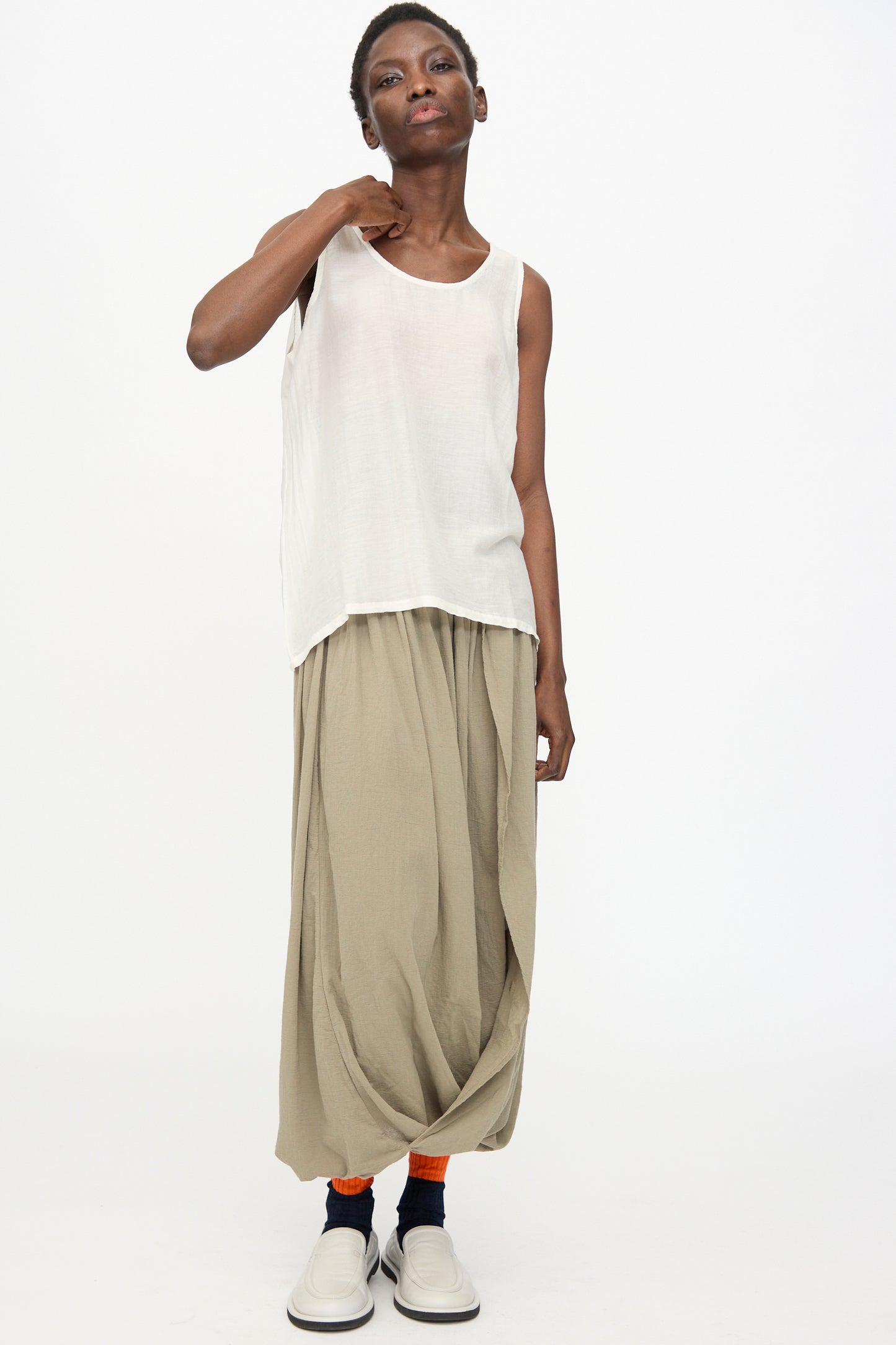 A person wears the Black Crane Loose Tank Top in White, beige draped pants, orange socks, and white slip-on shoes while standing against a plain white background.